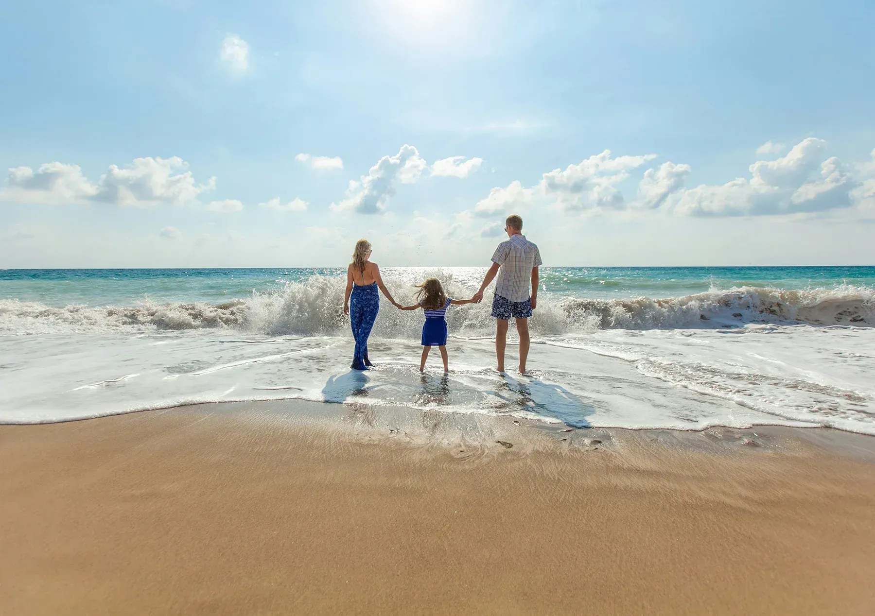 Three people holding hands at the beach as waves crash under a bright blue sky.