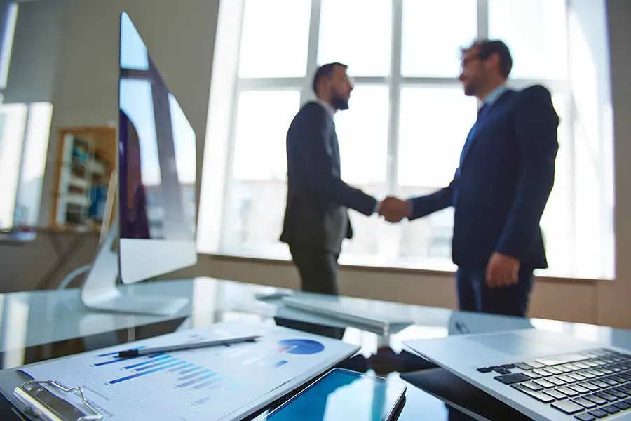 Two professionals shaking hands in a bright office with laptops on a conference table.