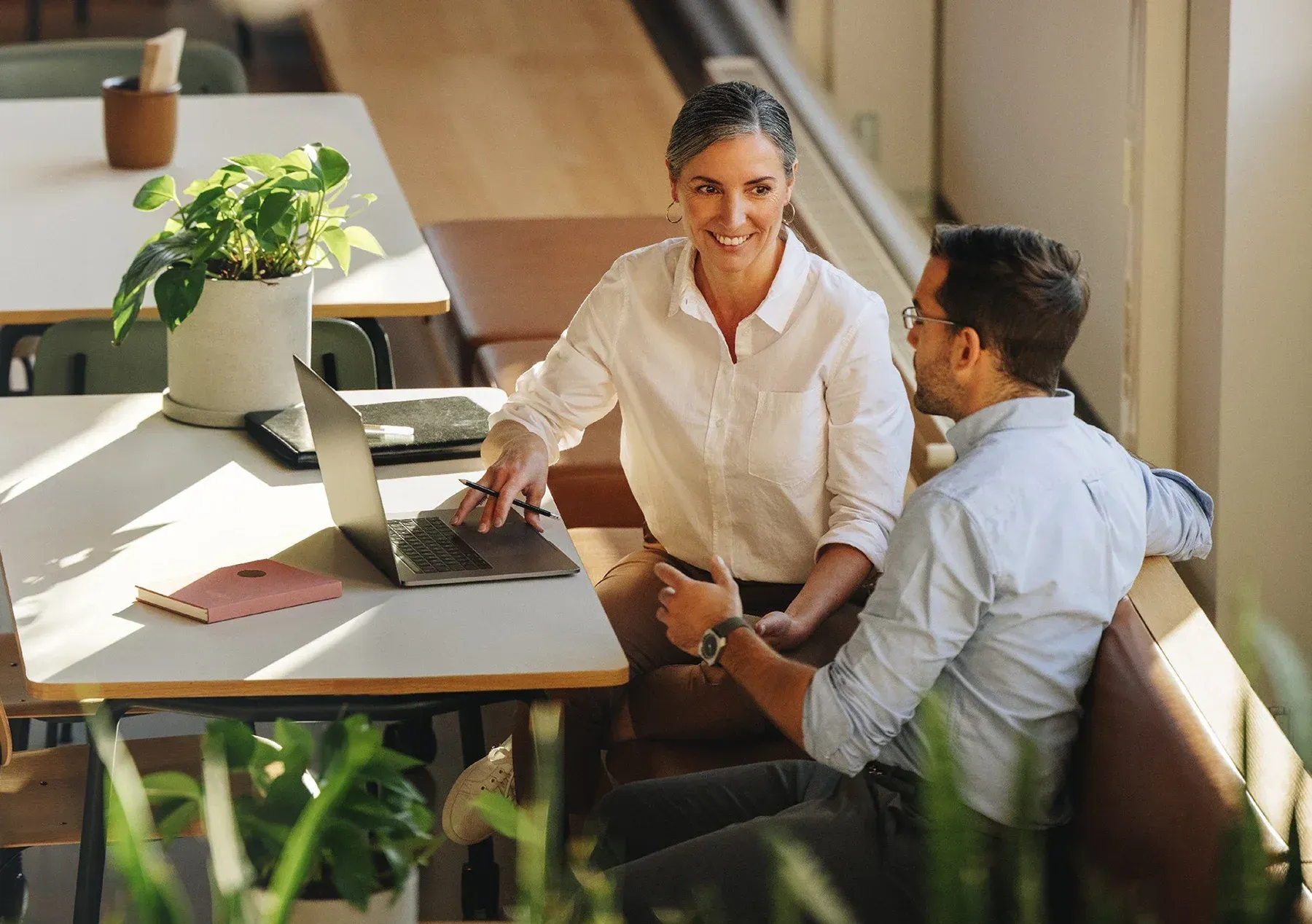 Two coworkers meeting at a table, smiling and using a laptop in a bright office.