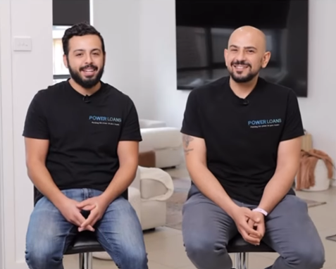Two smiling people in black T-shirts sit on stools in a bright office setting.