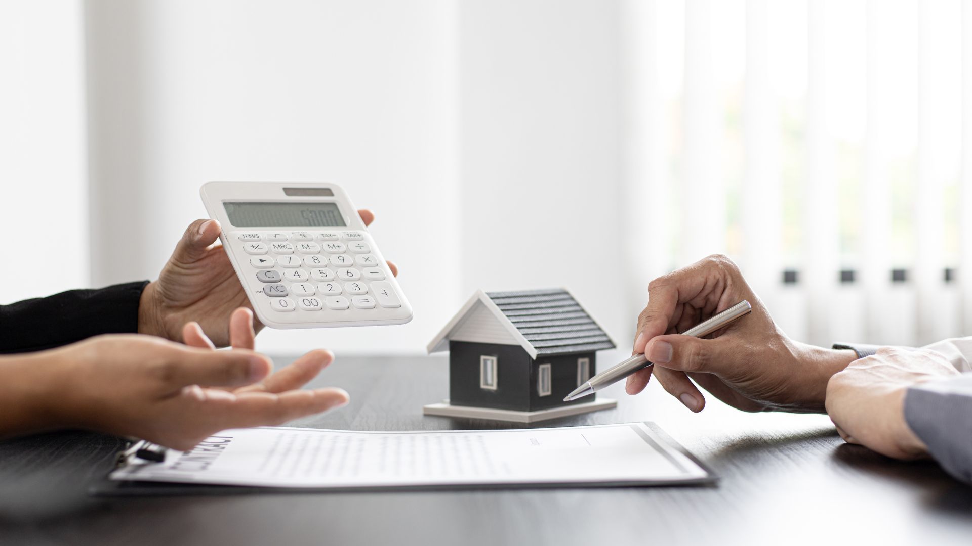 Hands discussing a house model and calculator over documents at a desk