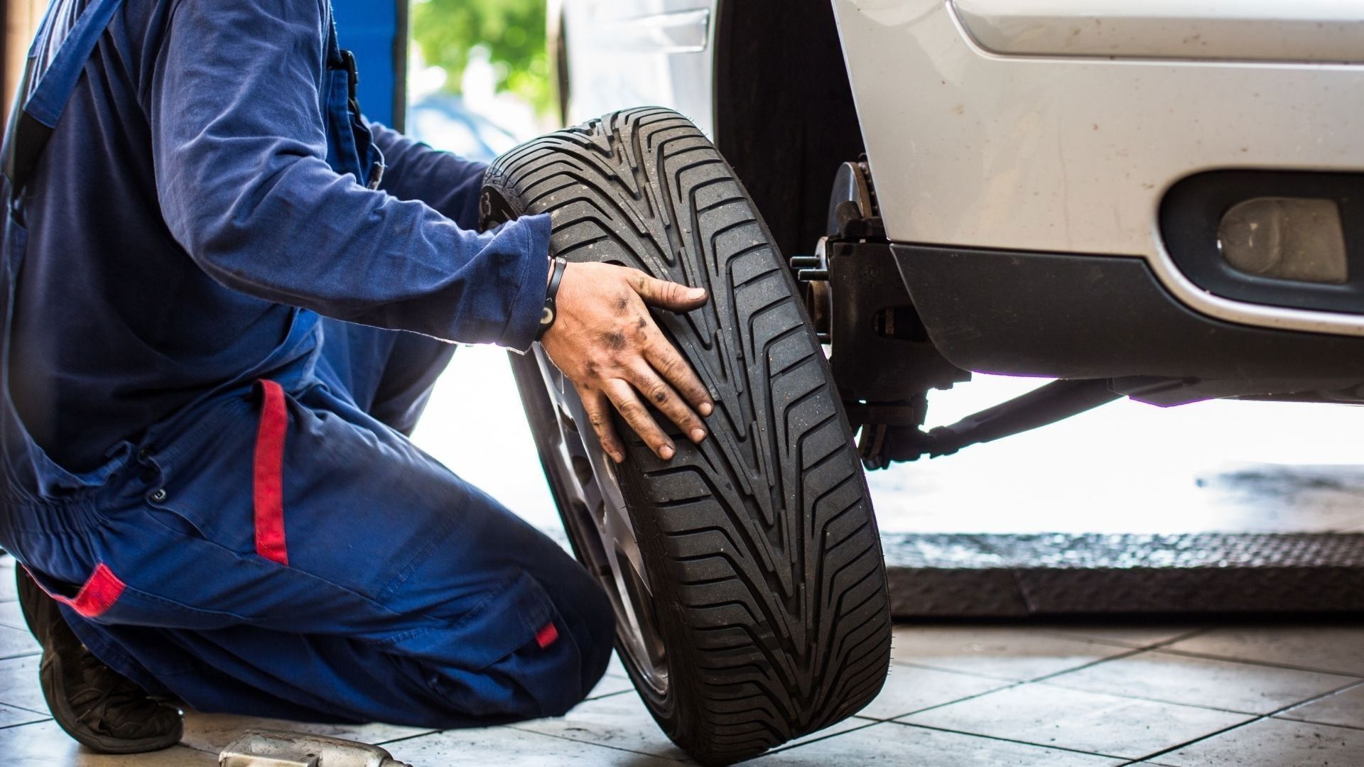 Mechanic kneeling beside a car, checking a detached tire in a garage.