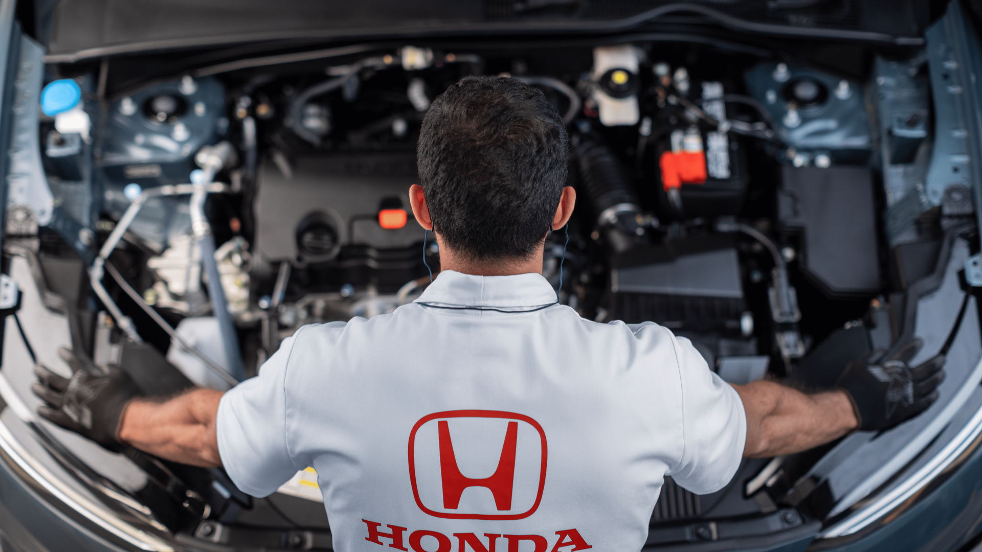Mechanic in white Honda shirt inspecting an open car engine bay from the rear