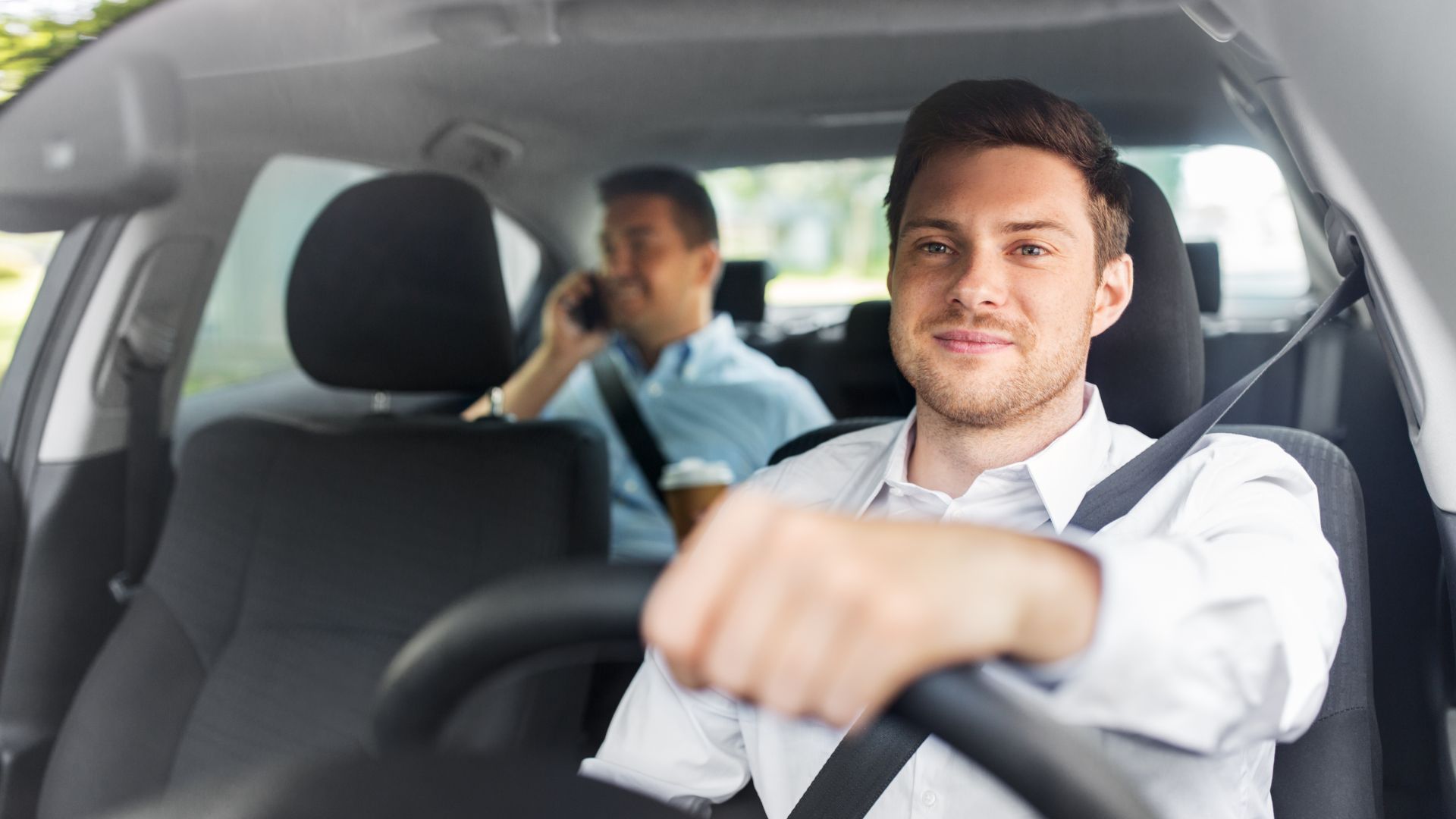 Driver smiling in the front seat of a car, with a passenger blurred in the back seat.