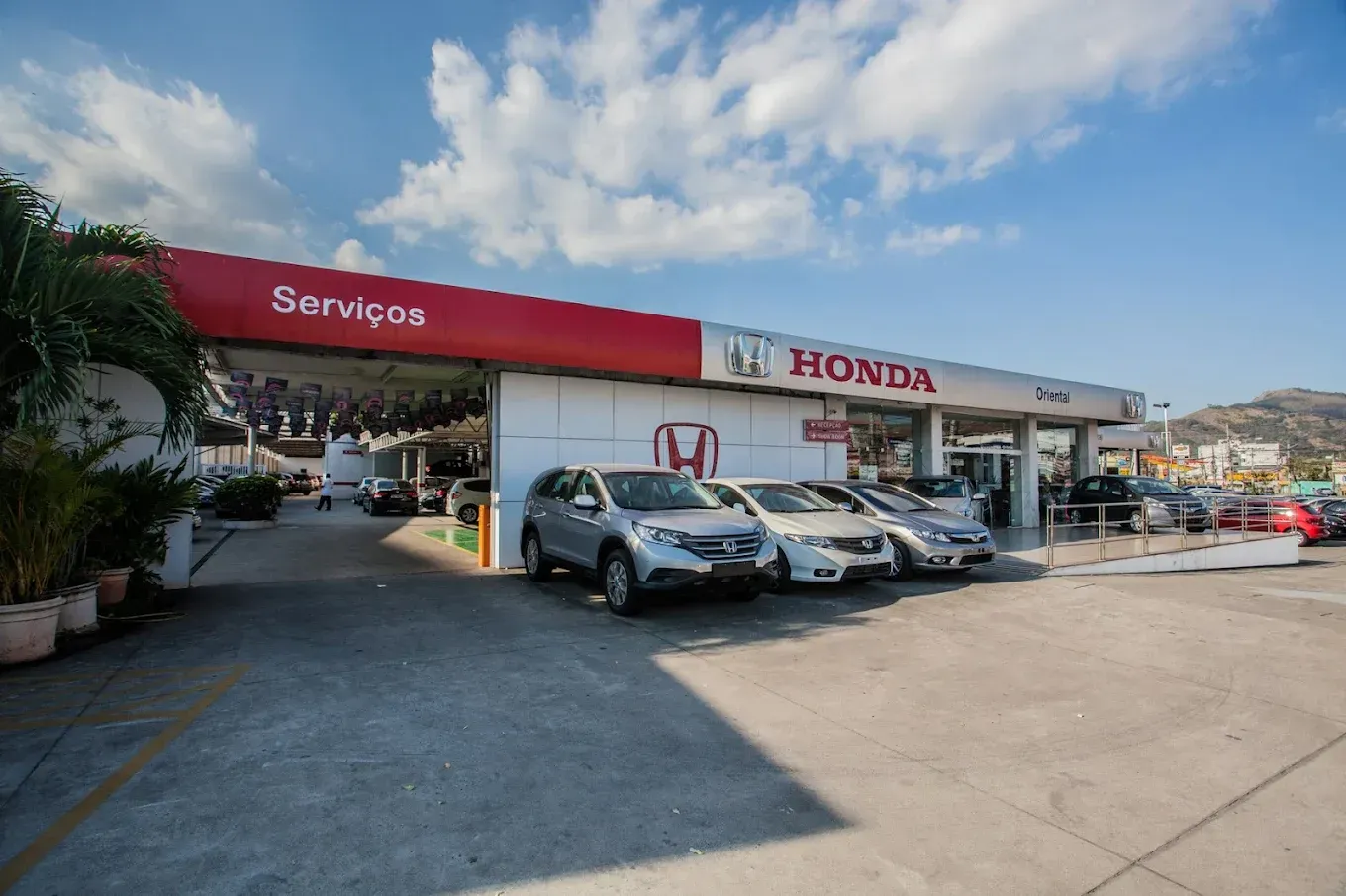Centro de serviço da Honda com carros estacionados do lado de fora, sob um toldo vermelho e céu azul.