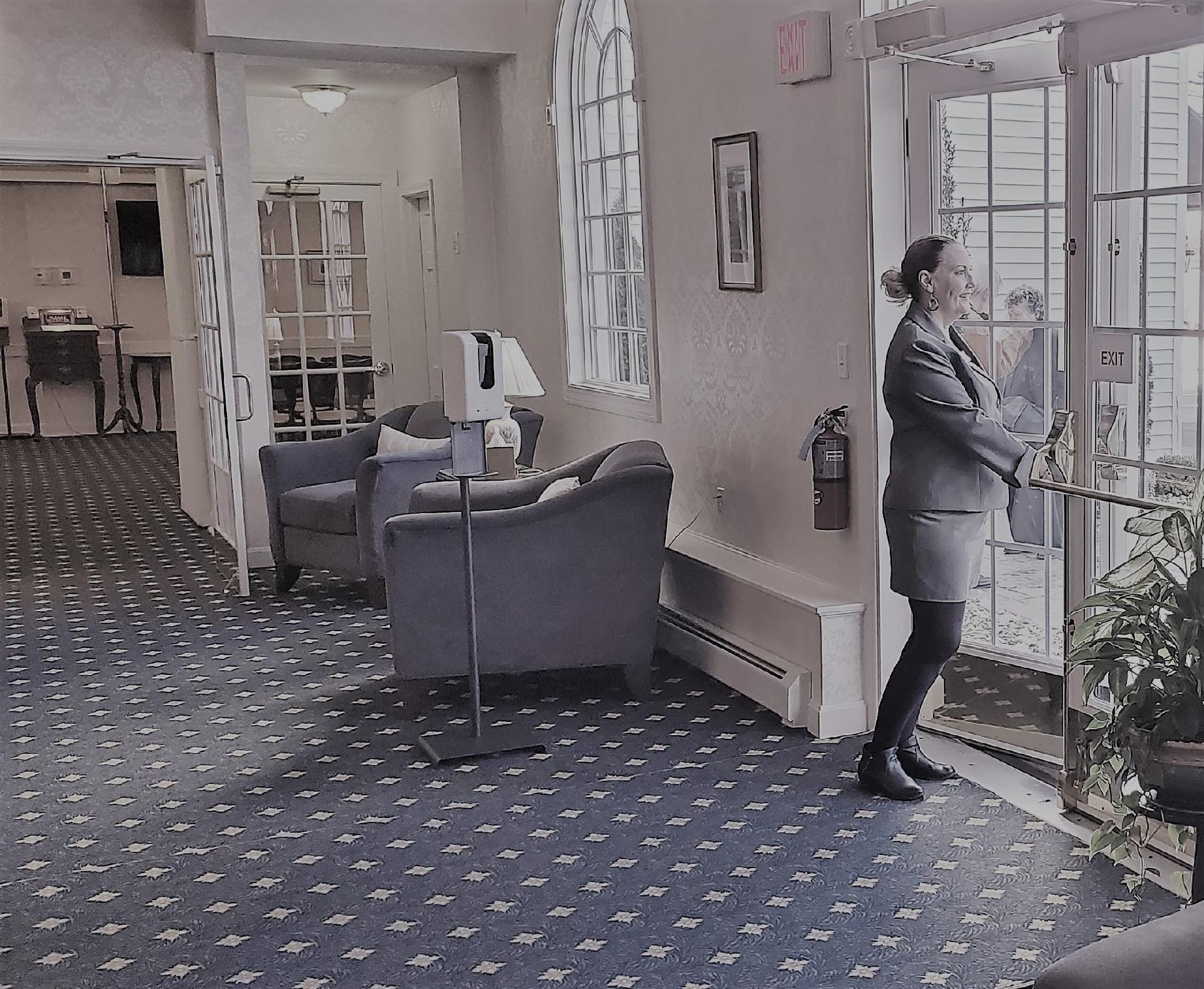 Lobby with blue chairs and patterned carpet; a person stands by glass doors, looking outside.