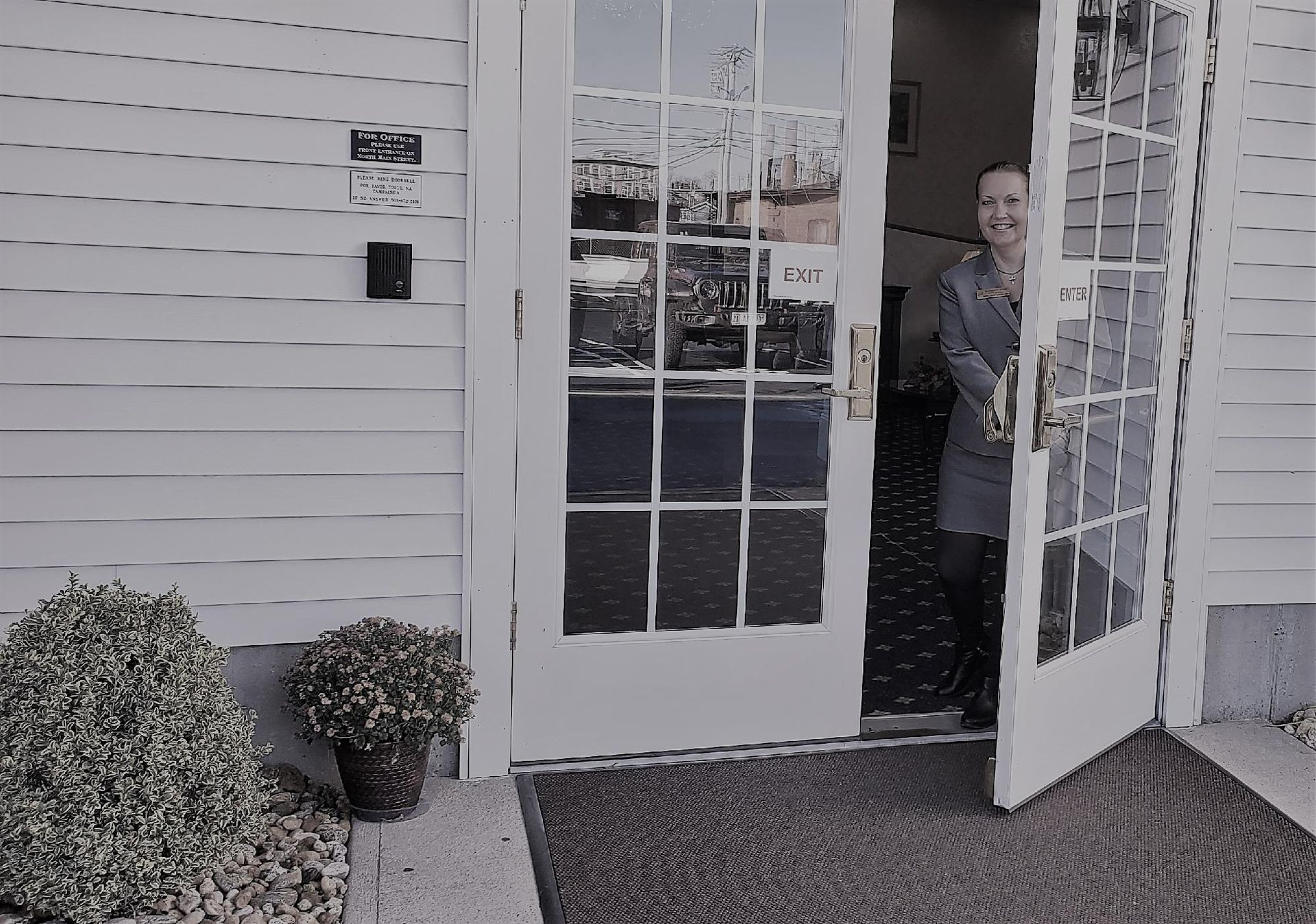 Person standing in a doorway of a white building, holding an open glass door.