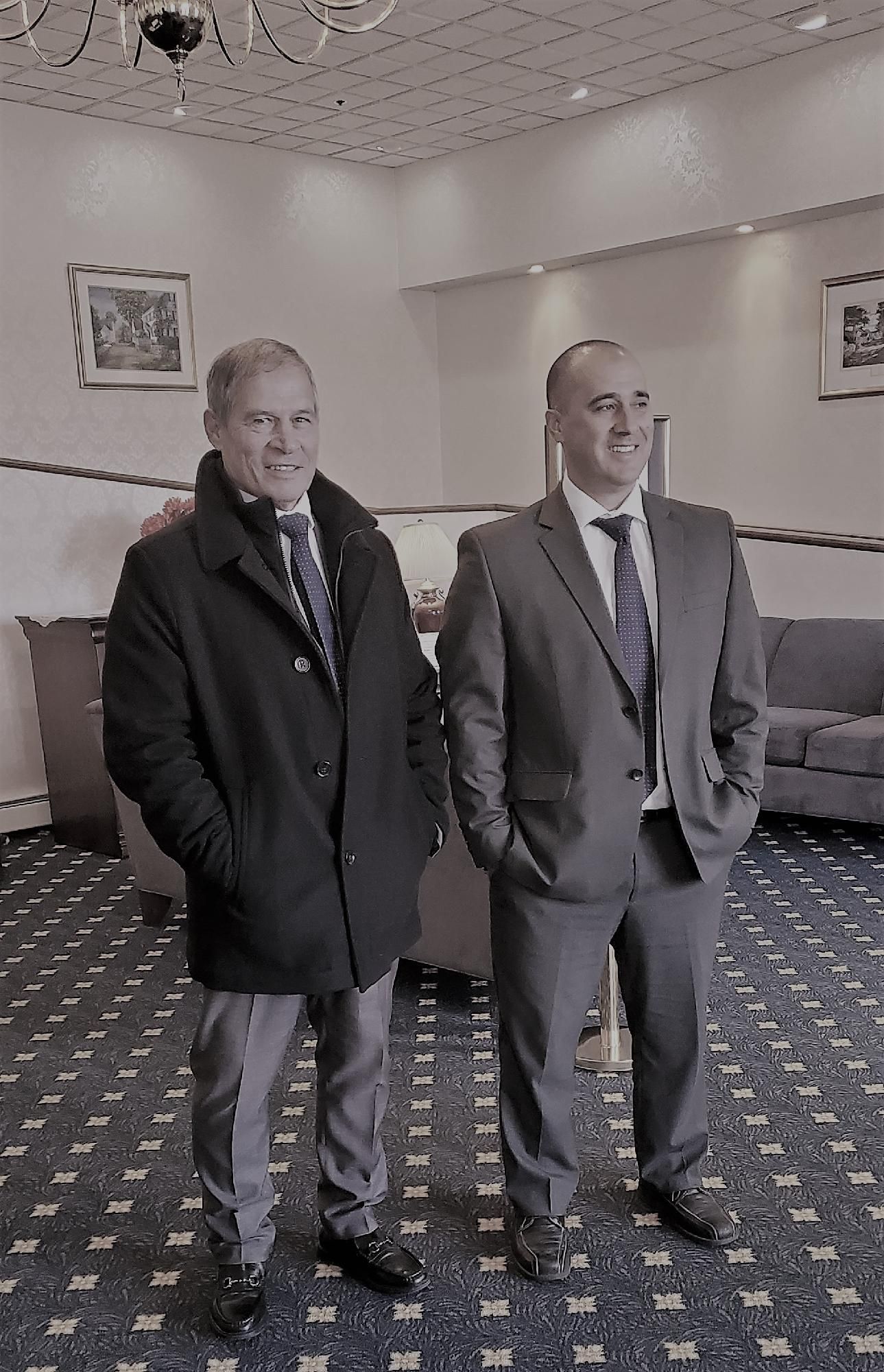 Two men in suits standing in a carpeted lounge, one with hands in pockets.