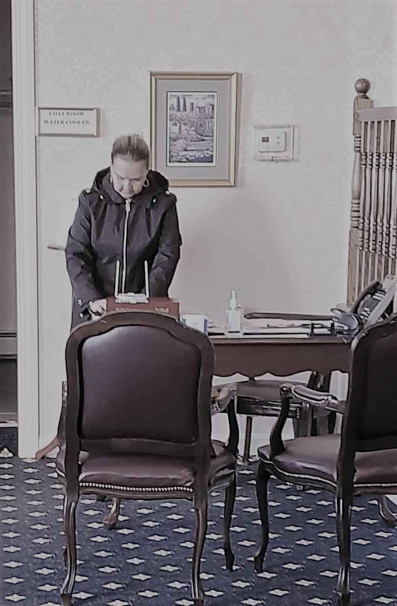 Person standing at a table in a room with chairs and patterned carpet, looking down at papers.