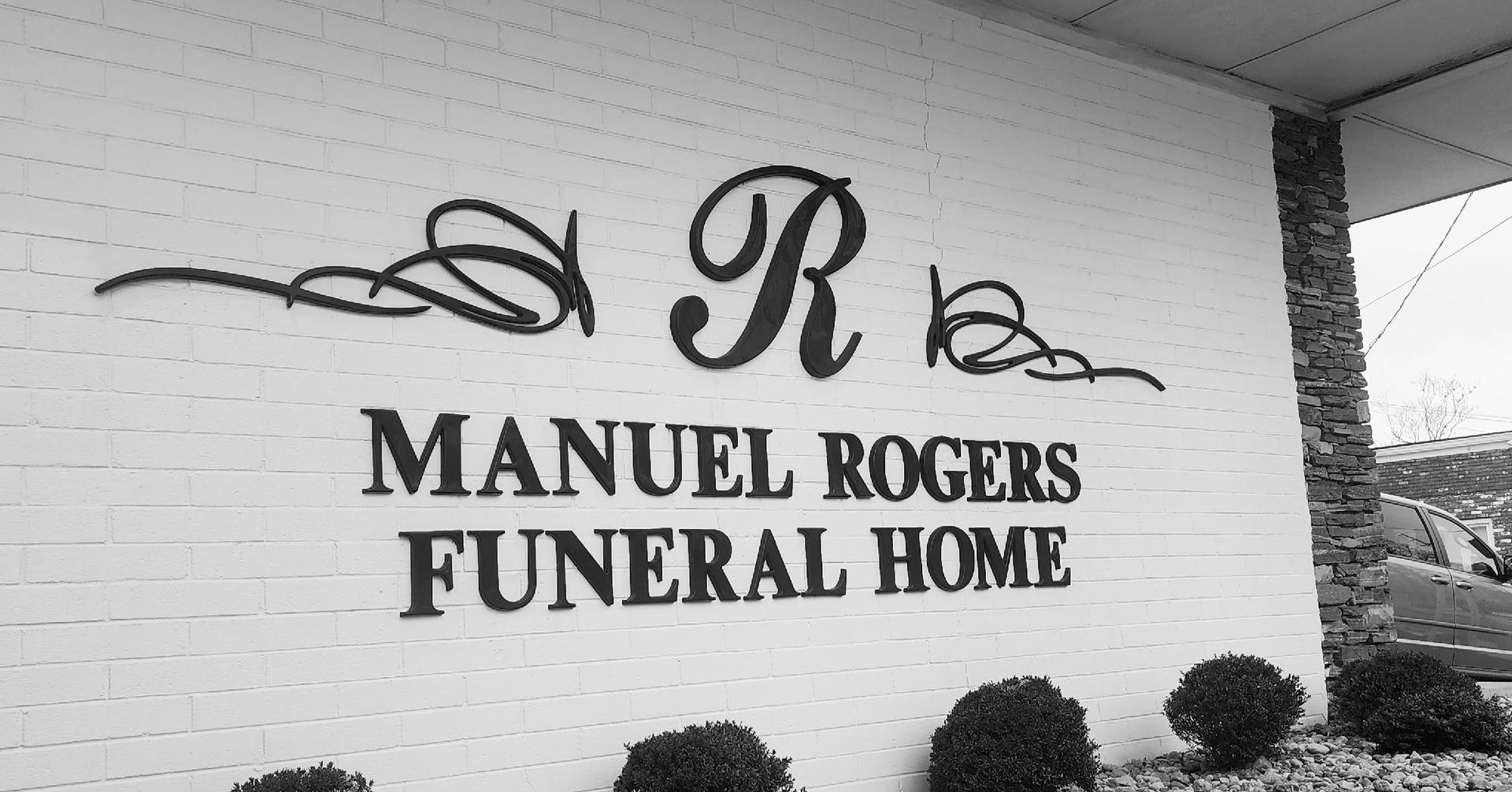 Manuel Rogers Funeral Home sign on a brick wall with decorative script and shrubbery below