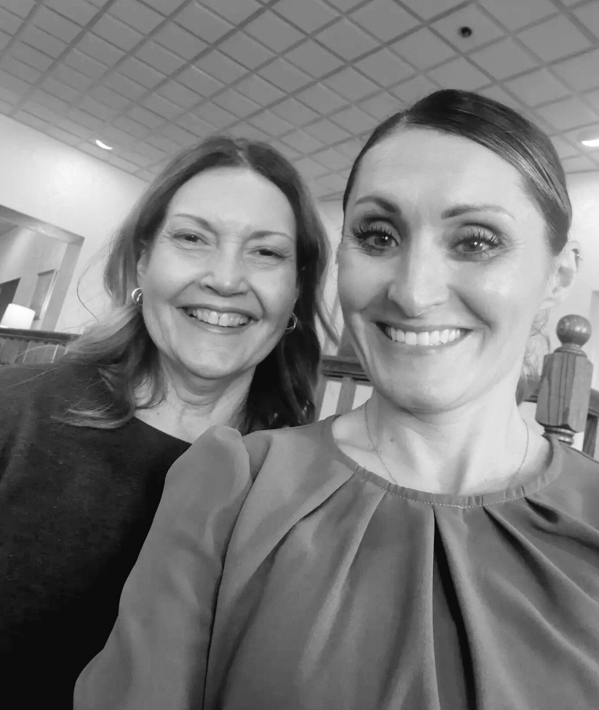 Two smiling women posing for a selfie indoors under a tiled ceiling.