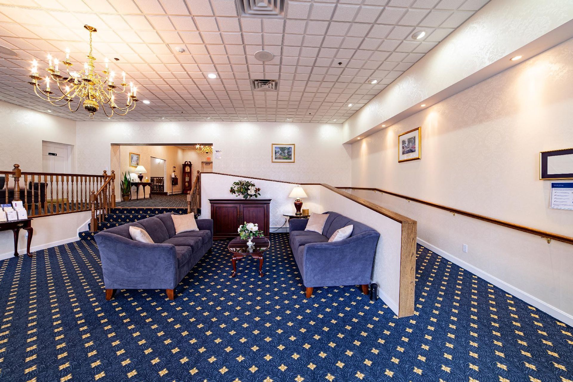 Spacious hotel lobby with blue patterned carpet, chandeliers, and gray armchairs around a small table.
