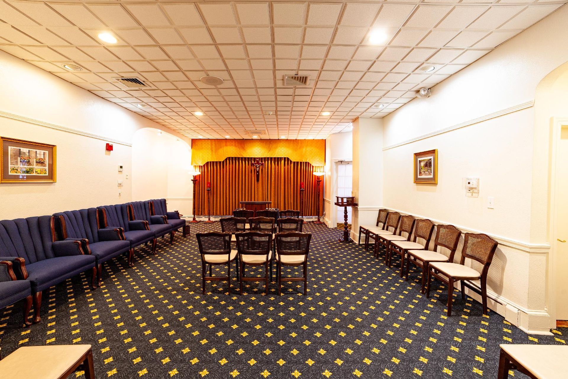Empty meeting room with rows of chairs, patterned carpet, and a small stage with orange curtains.