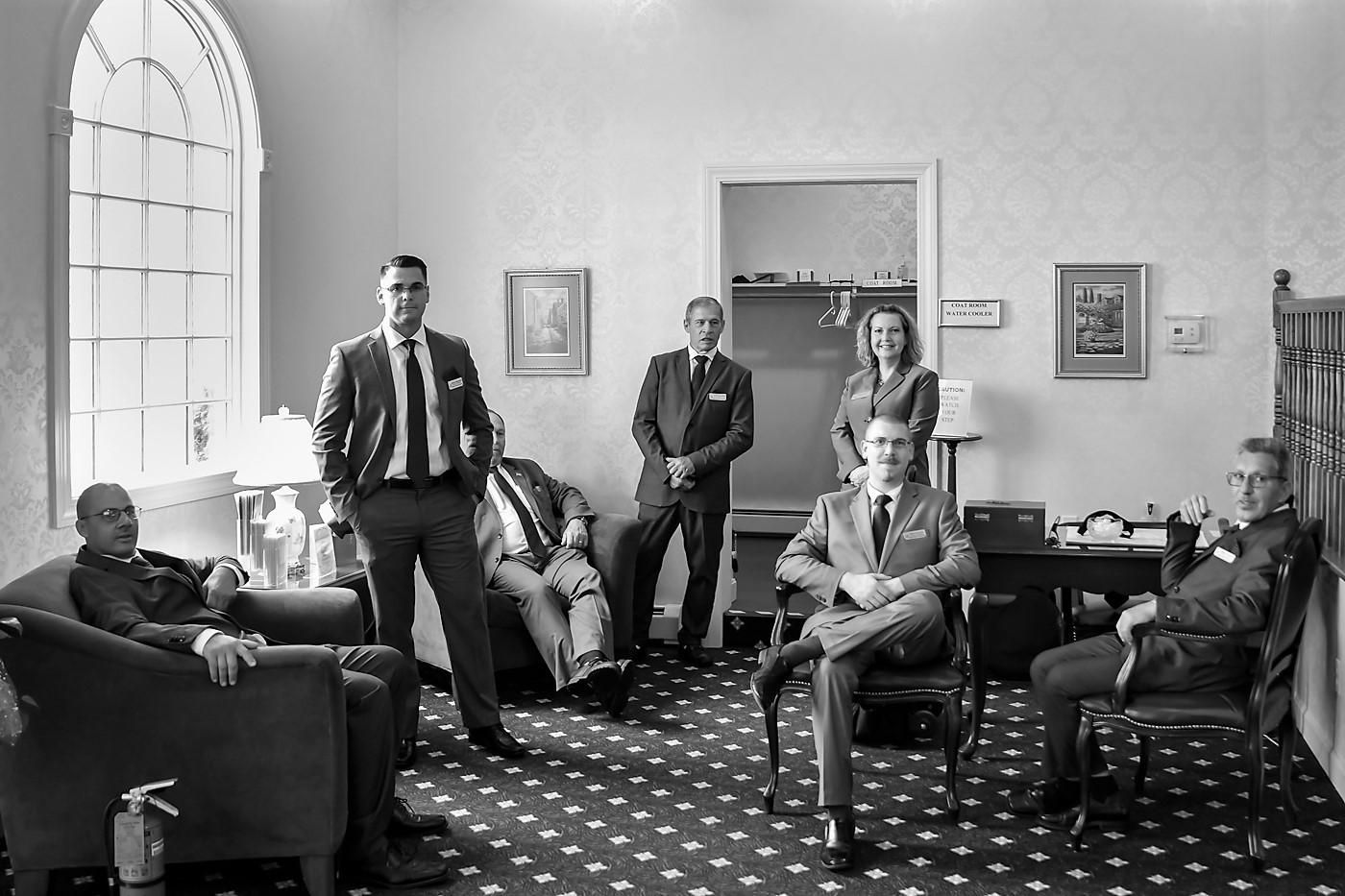 Group of people seated and standing in a bright parlor with patterned carpet and large arched window.