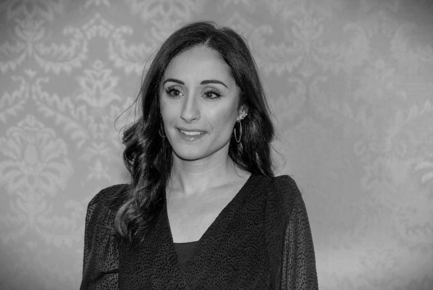 Woman in a dark top standing before patterned wallpaper, smiling slightly at the camera.