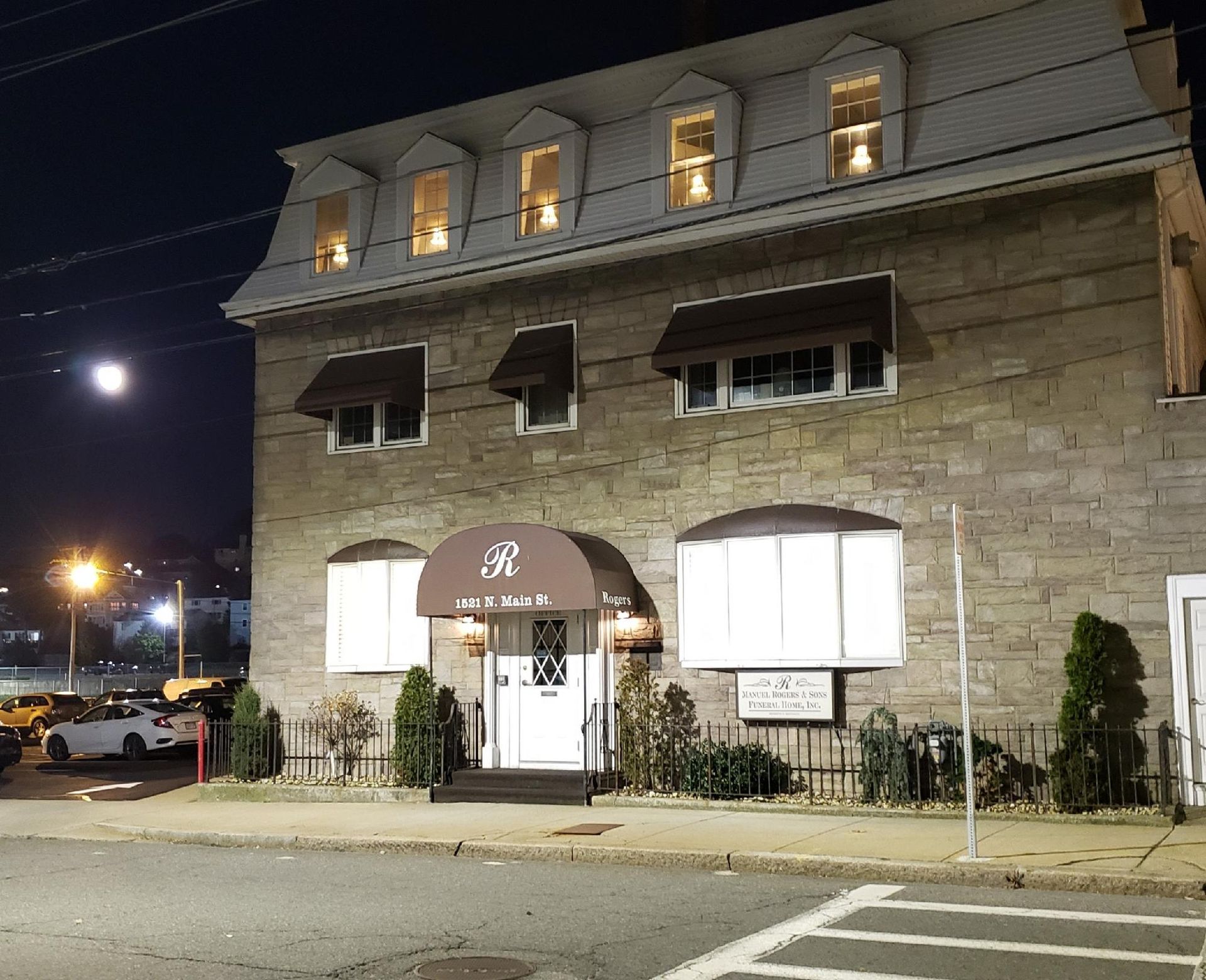 Nighttime view of a stone building with lit windows, awnings, and a glowing entrance sign.