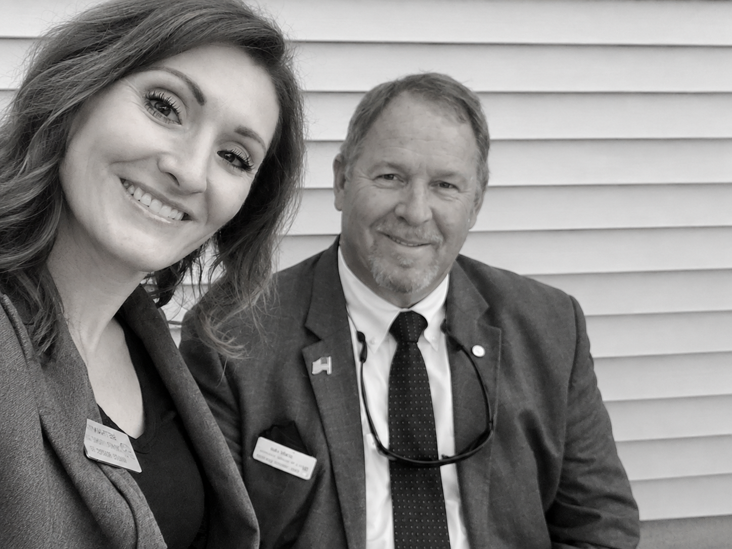Two people smiling in suits, seated outdoors in front of horizontal siding.