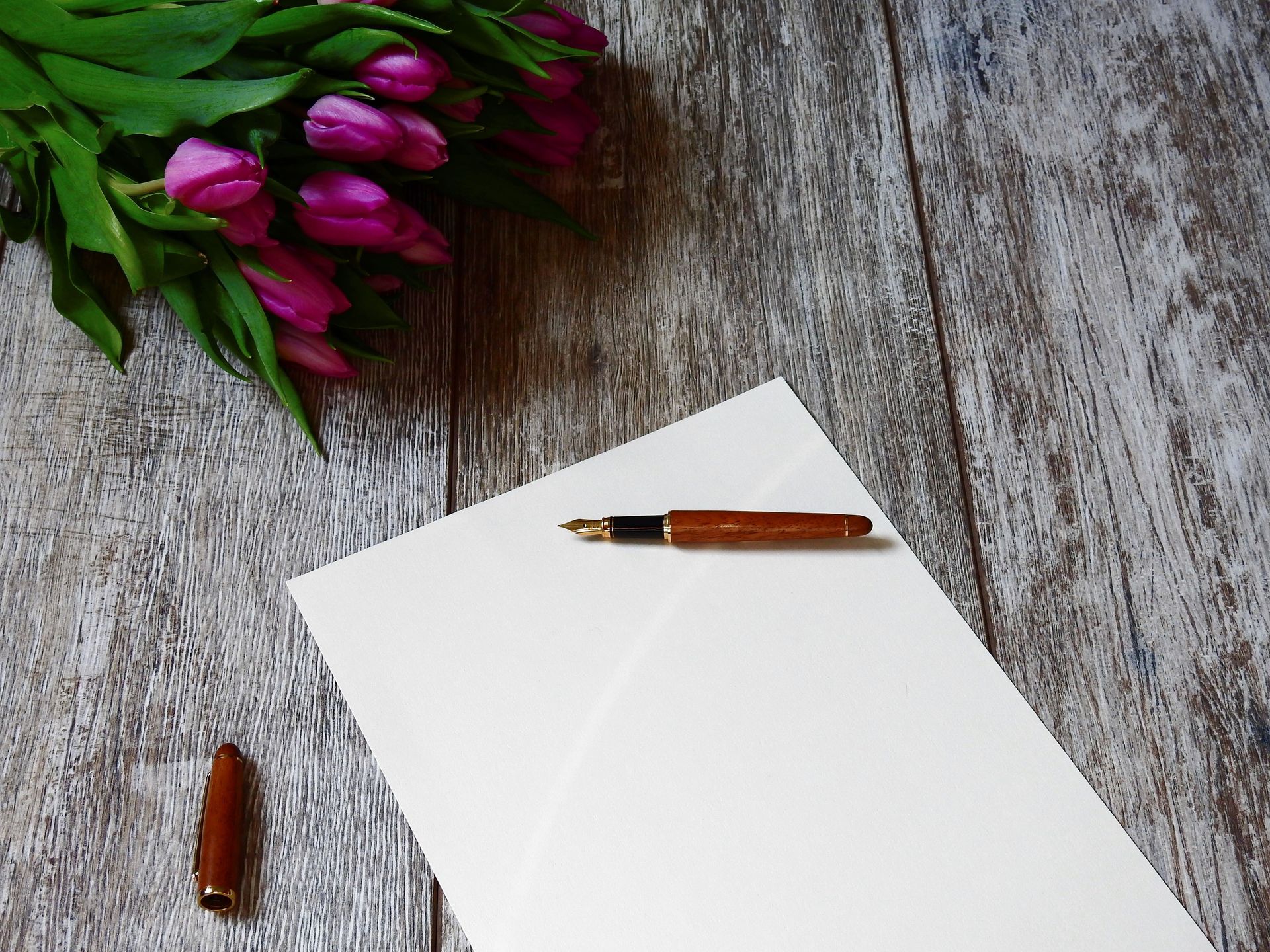 White paper with two fountain pens on a rustic wooden table, beside pink tulips and leaves.