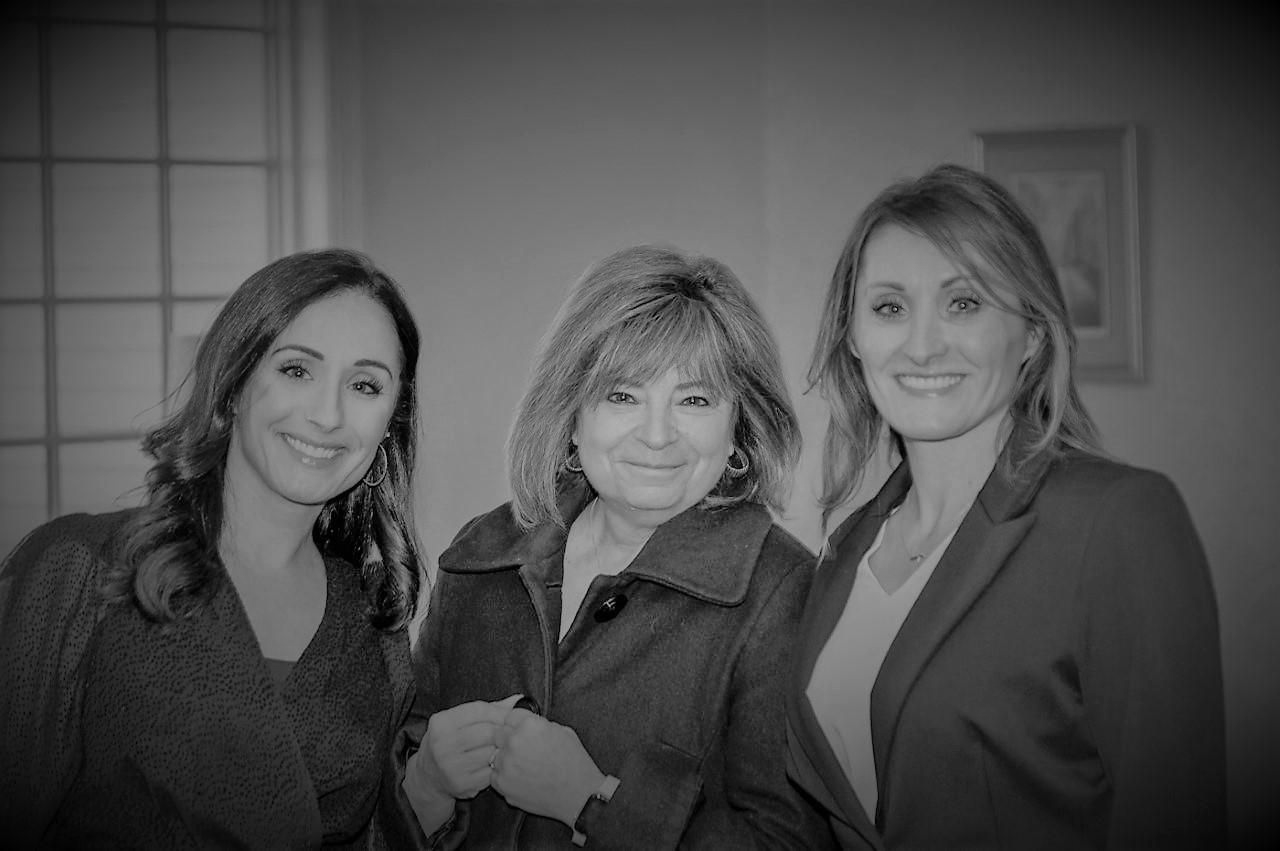 Three women in business attire smiling indoors, standing together in an office setting.