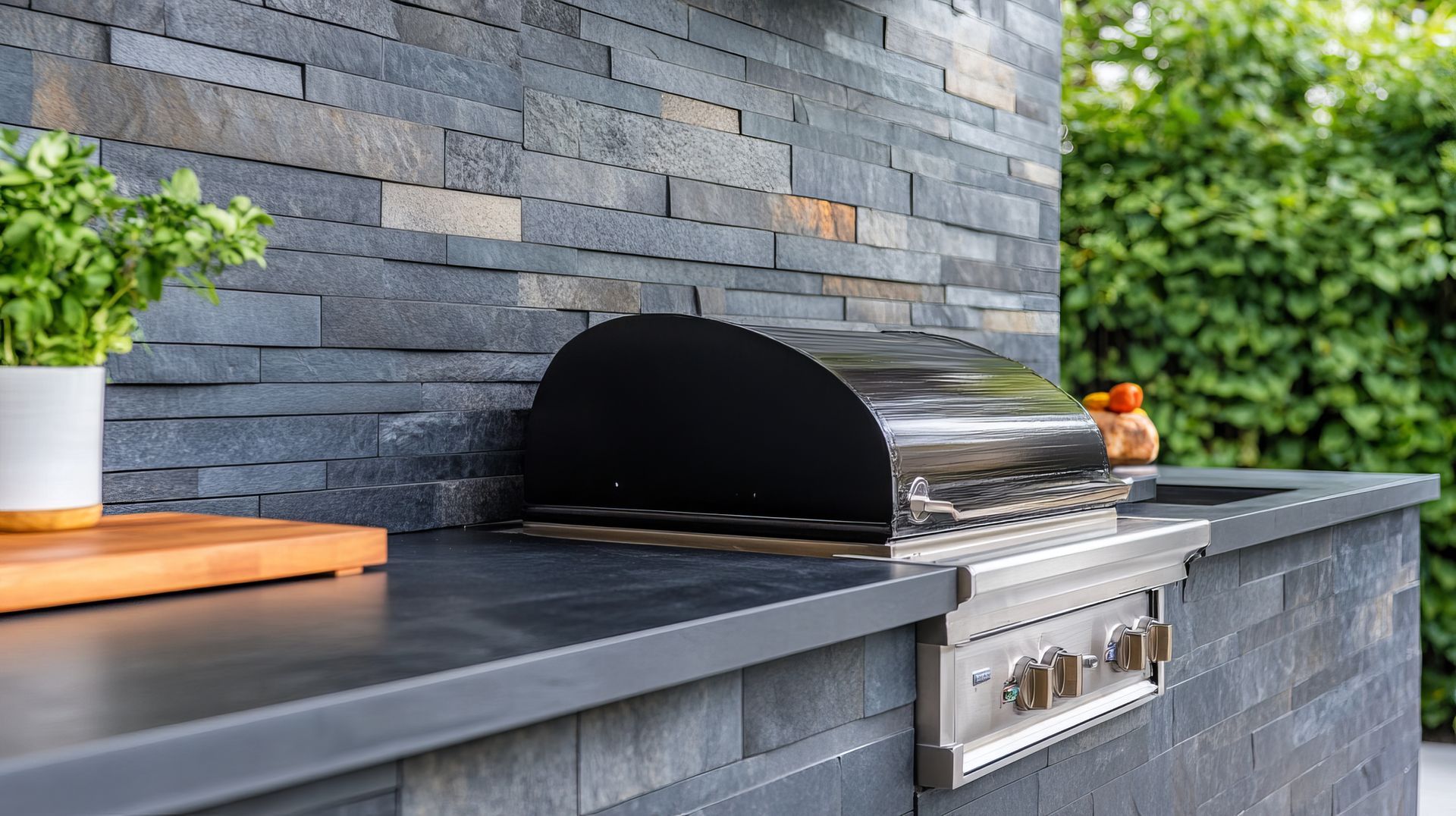 Une cuisine extérieure avec un barbecue en acier inoxydable, un plan de travail en pierre noire et un mur carrelé, agrémentée d'une petite plante en pot.