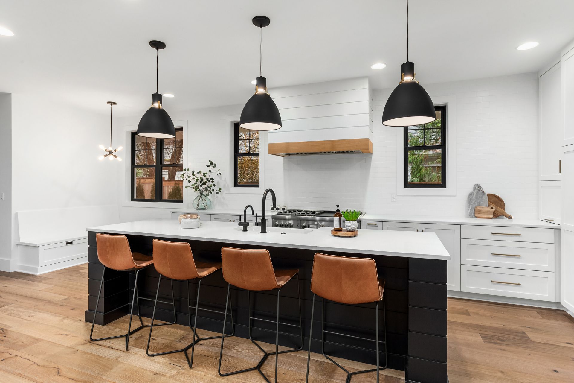 Une cuisine moderne avec un îlot central noir et quatre tabourets en cuir beige, éclairés par trois suspensions noires.