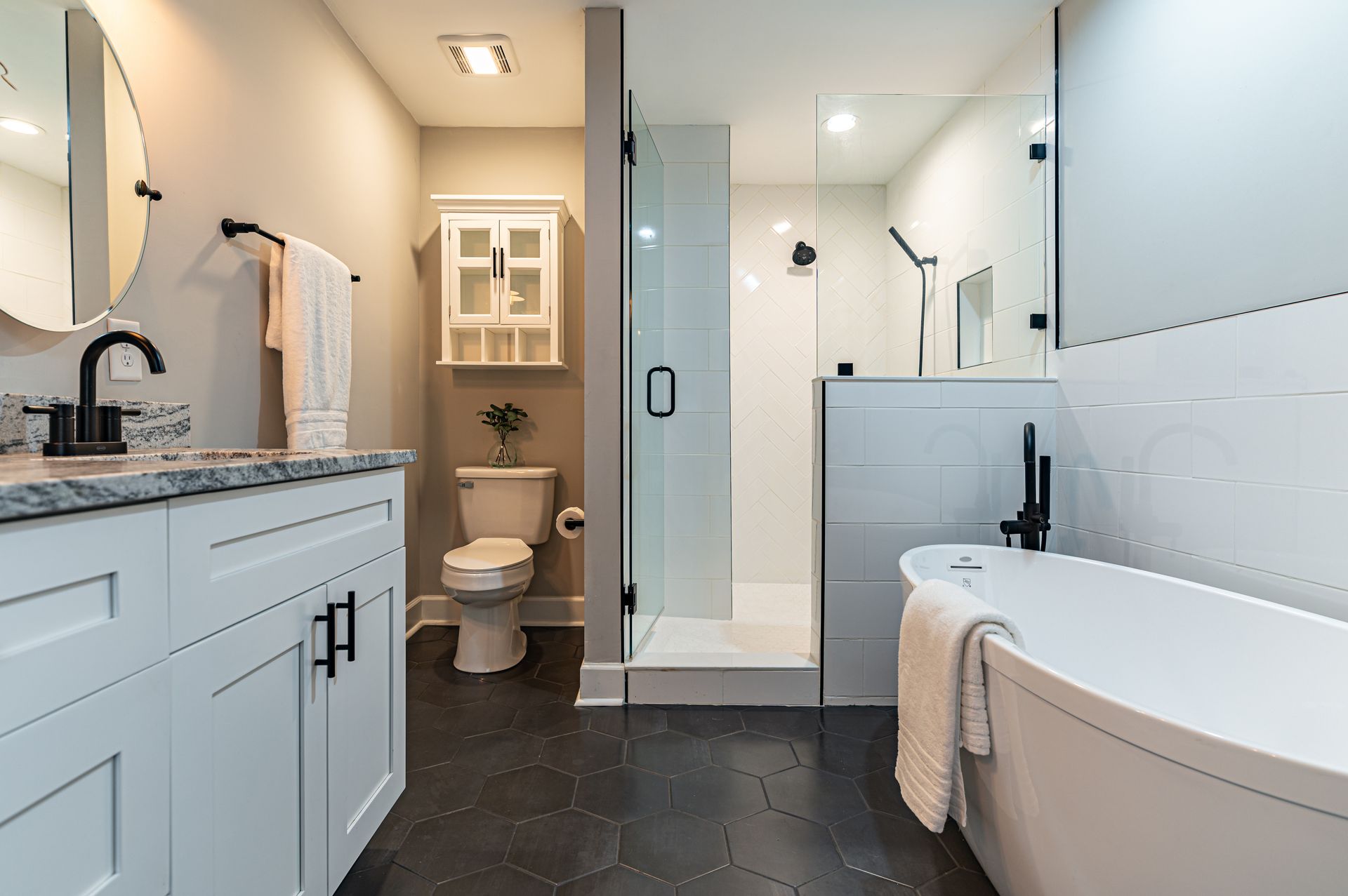 Salle de bains moderne avec meuble-lavabo blanc, baignoire îlot, douche vitrée et toilettes sur un sol carrelé de carreaux hexagonaux noirs.