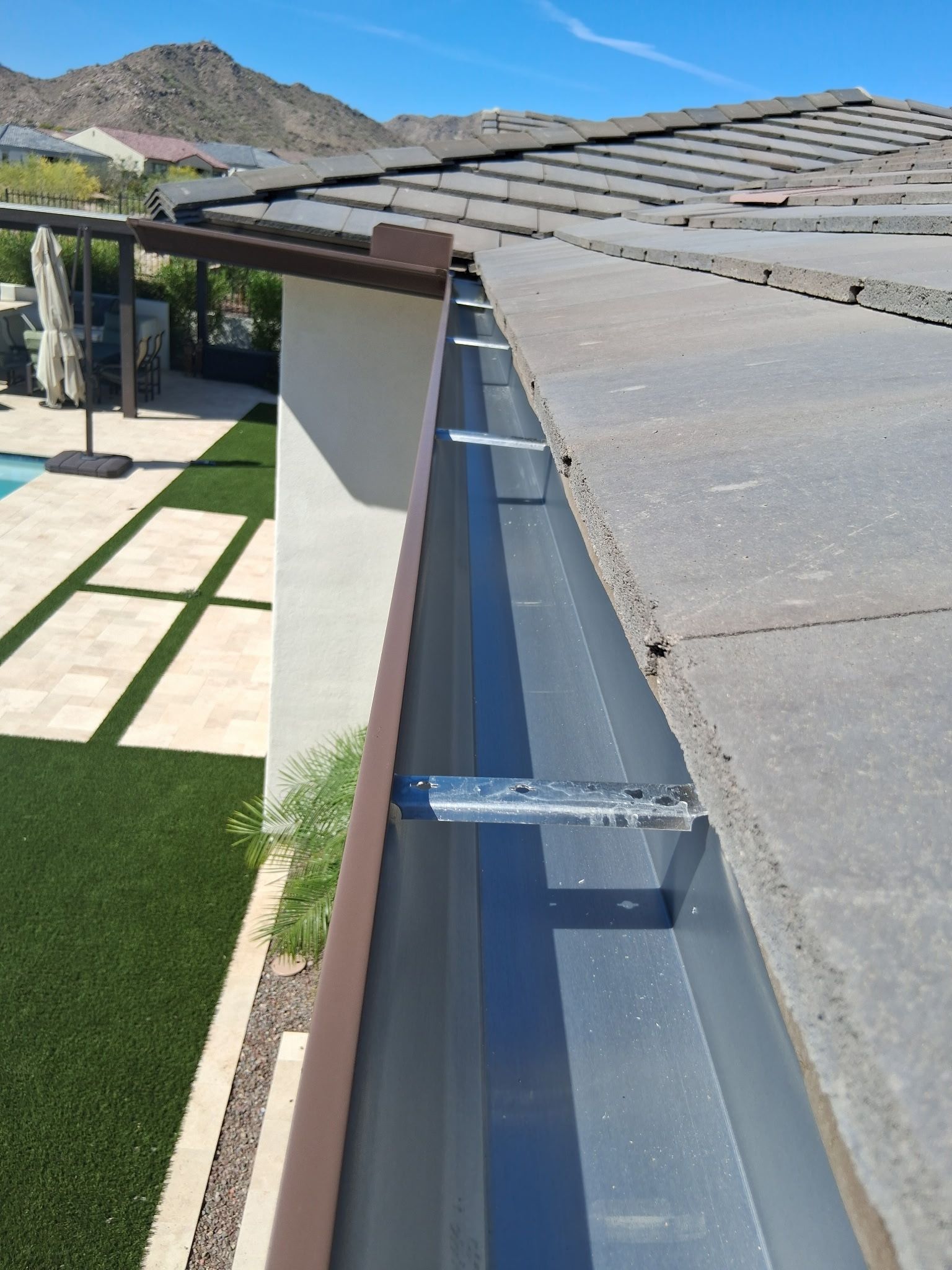 A view looking down along a metal rain gutter installed on a tile roof, with a patio and lawn visible below.