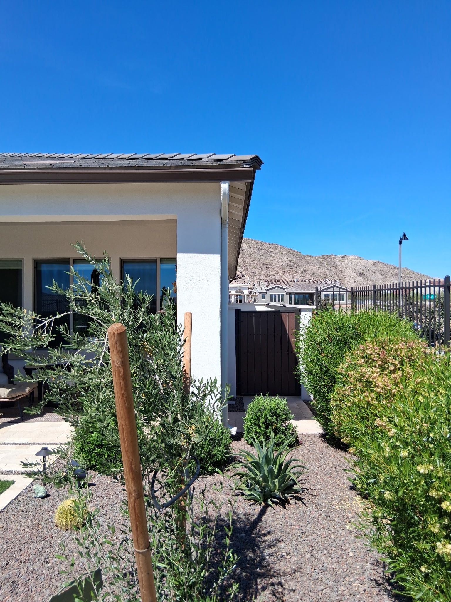 A view of a house exterior with a side patio, landscaped yard, and a wooden gate, framed by a bright blue desert sky.