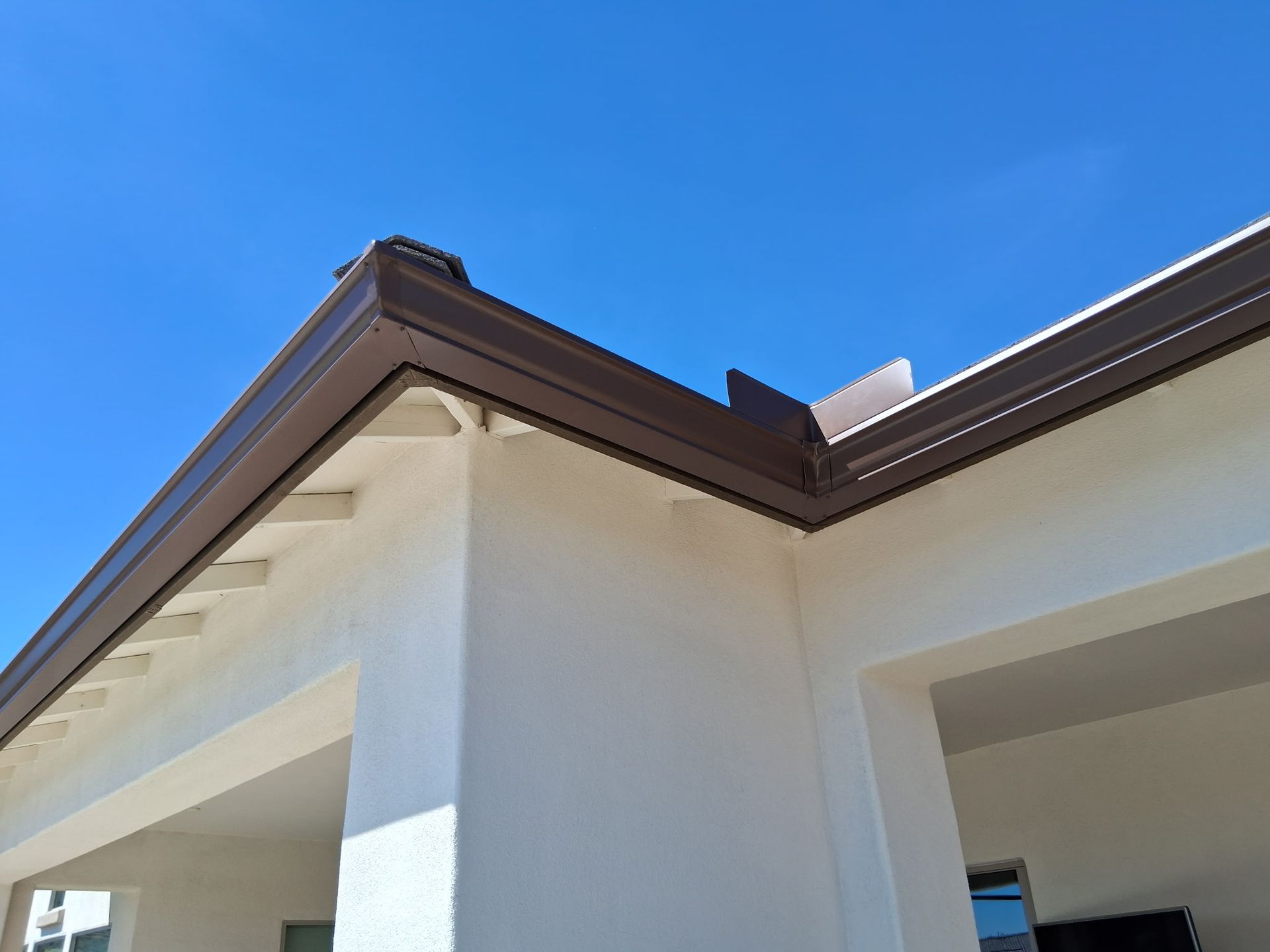 A low-angle view of a white stucco house corner with brown rain gutters against a bright blue sky.