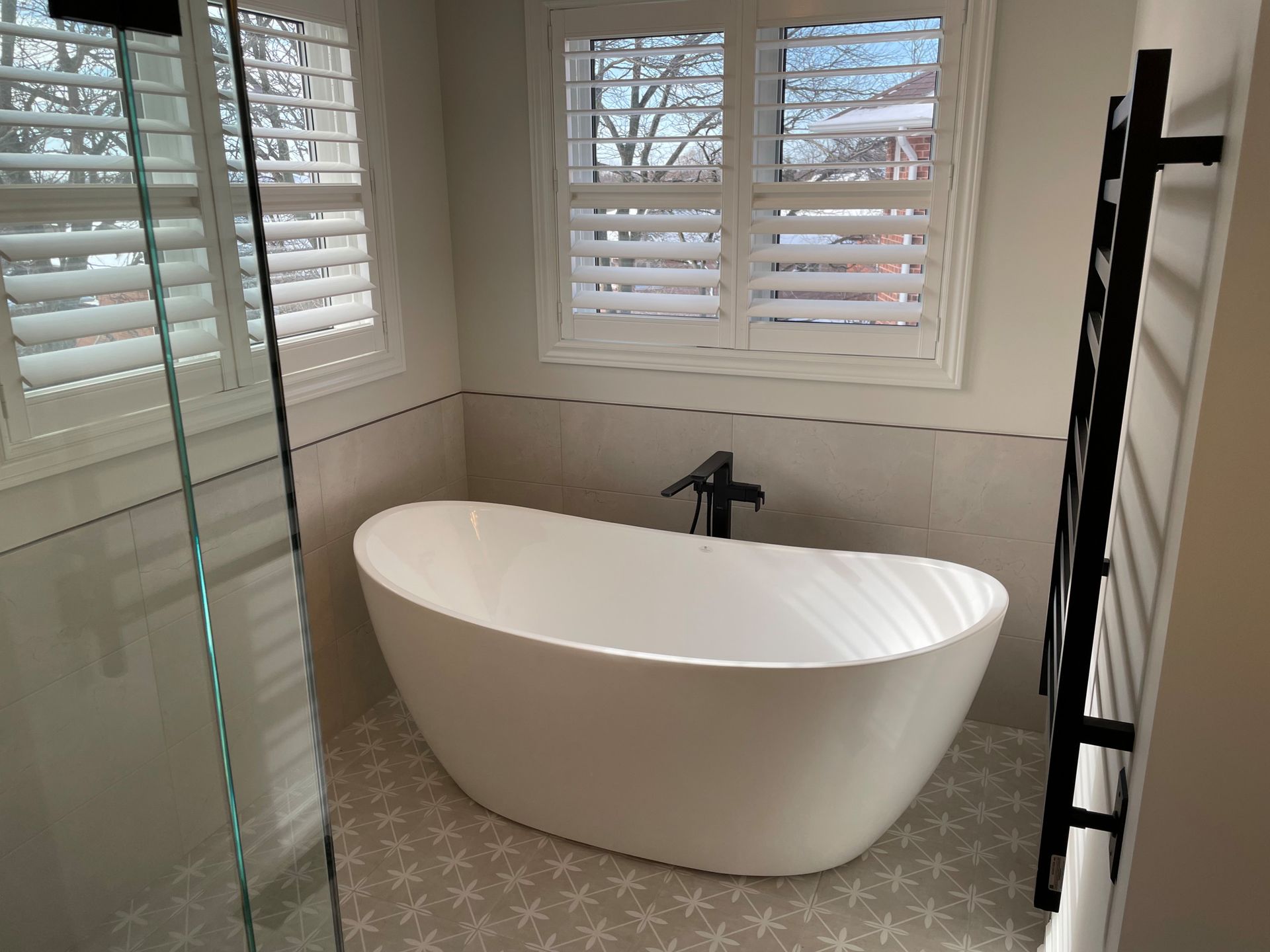 Modern bathroom with white freestanding tub, patterned tile floor, and window shutters.