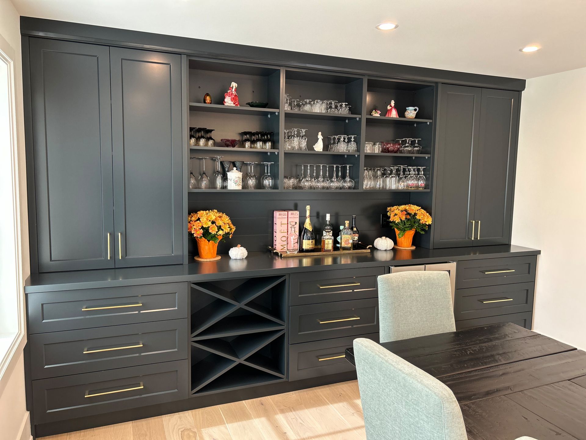 Dark gray built-in cabinet with wine rack and glass shelves, holding drinks and decor, next to a dining table.