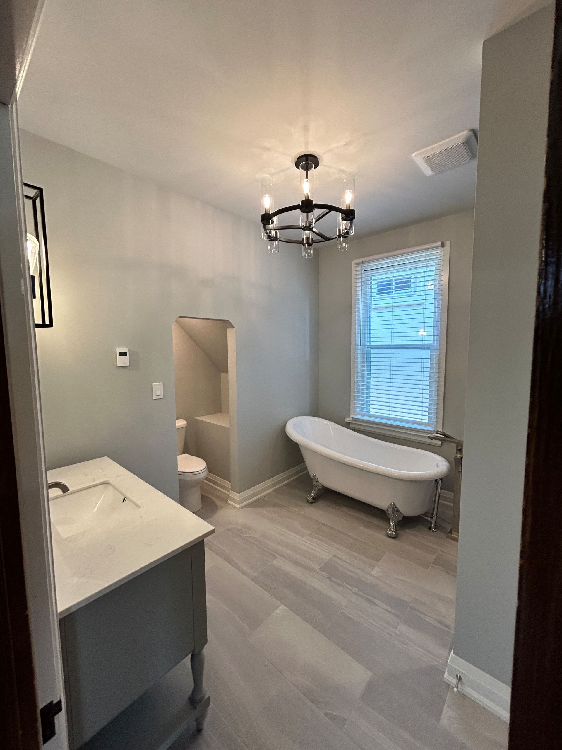 Bathroom with clawfoot tub, vanity, toilet, chandelier, and light gray walls and flooring.