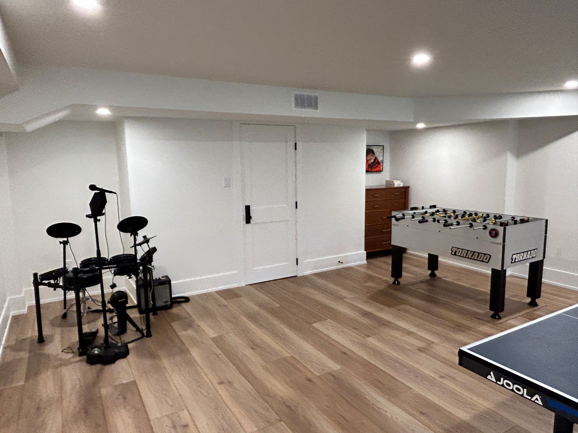 Game room with foosball table, drum set, and ping pong table; hardwood floors, white walls.