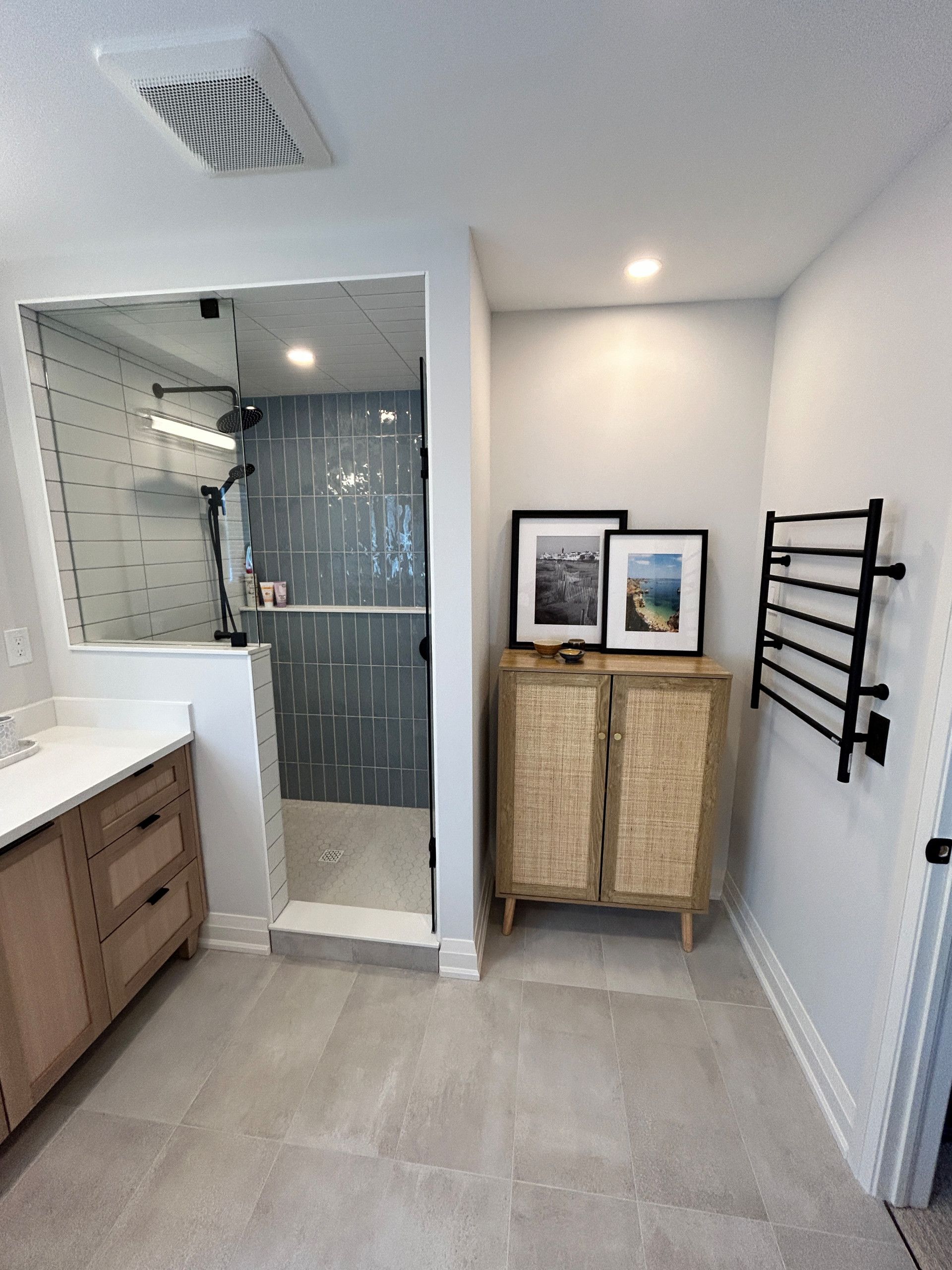 Bathroom with walk-in shower, light wood vanity, cabinet, black towel warmer, and framed photos.