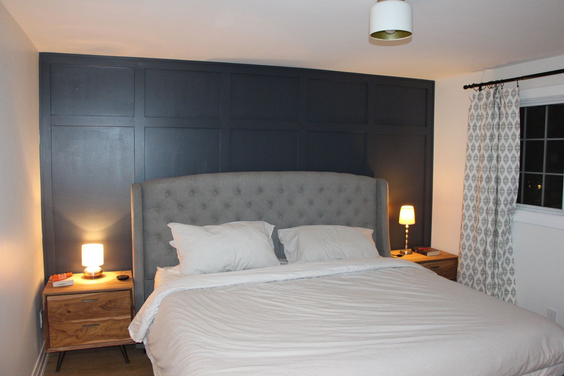 Bedroom with gray tufted headboard, nightstands, dark blue paneling on wall, and patterned curtains.