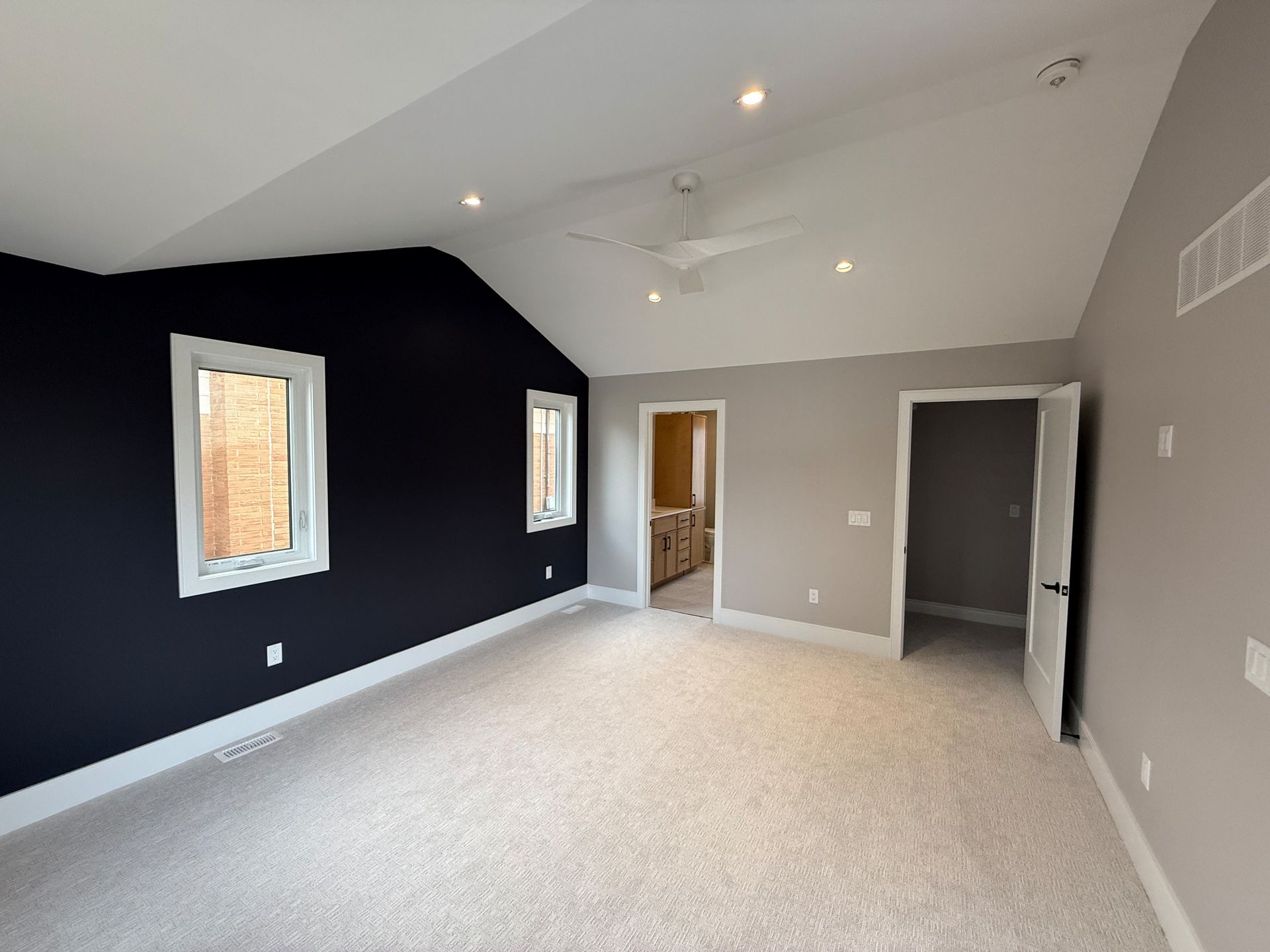 Empty bedroom with navy accent wall, beige walls, and white trim. Beige carpet, recessed lighting, and open doors.