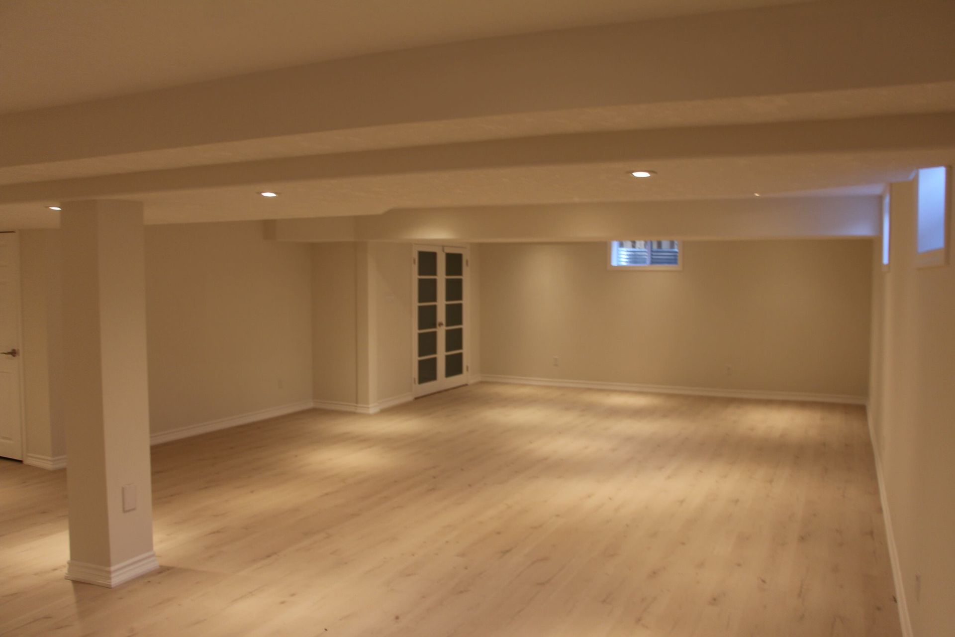 Empty, well-lit basement with light wood flooring, white walls, support beams, and a built-in cabinet.