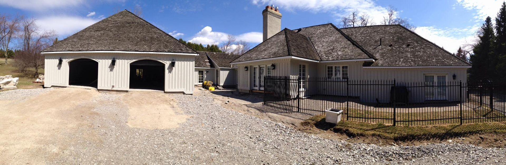 A light-colored house and garage with dark roofs on a gravel driveway under a blue sky.