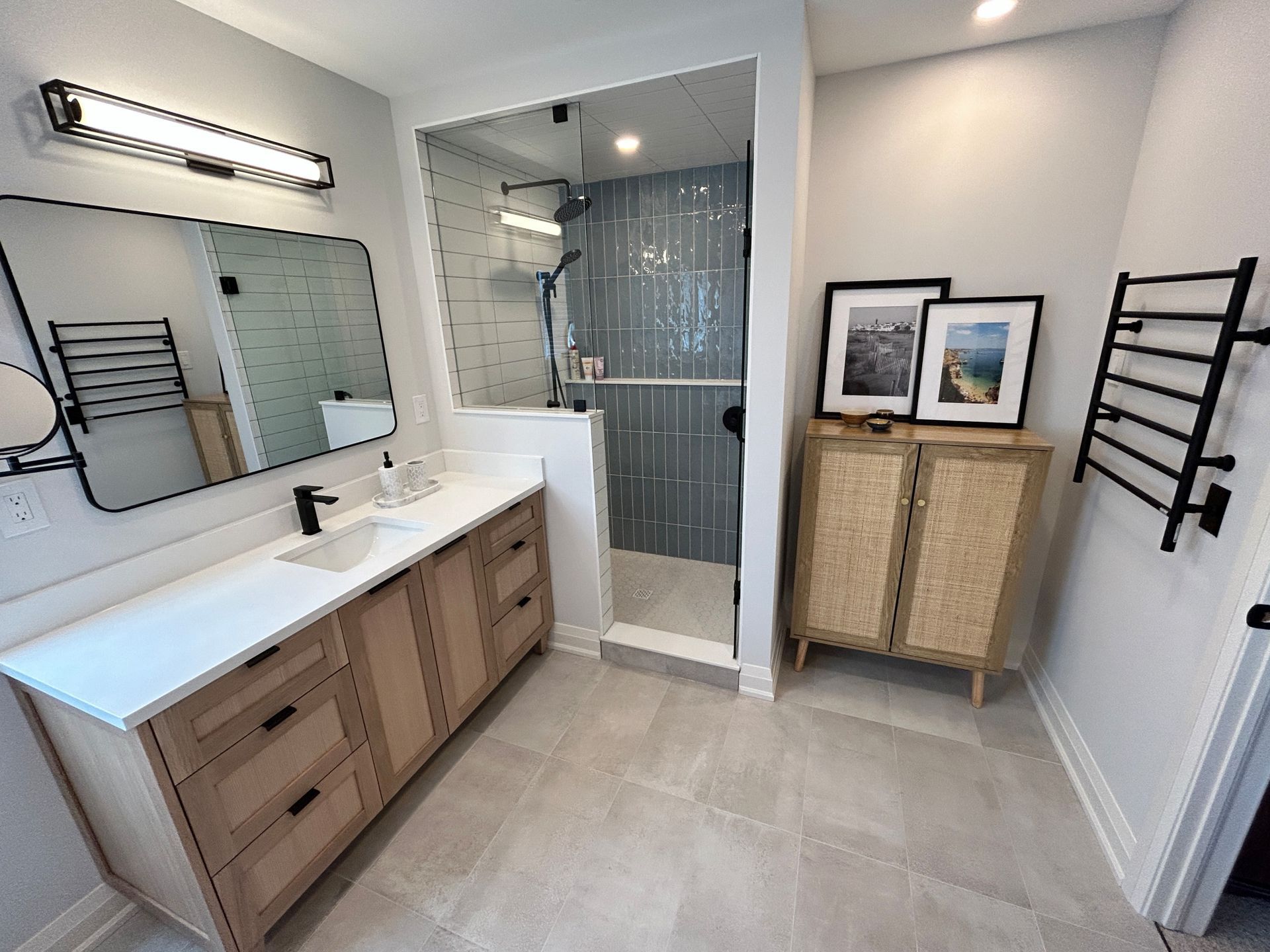 Modern bathroom: light wood vanity, gray tile floor, glass shower, decorative cabinet, black towel rack.