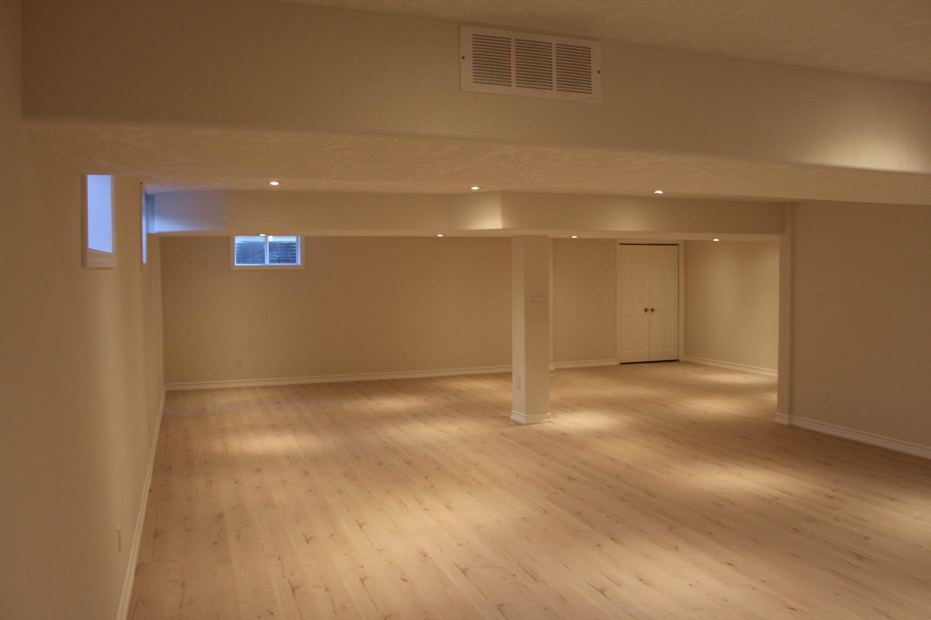 Empty basement room with light-colored wooden floor, tan walls, recessed lights, and support beams.