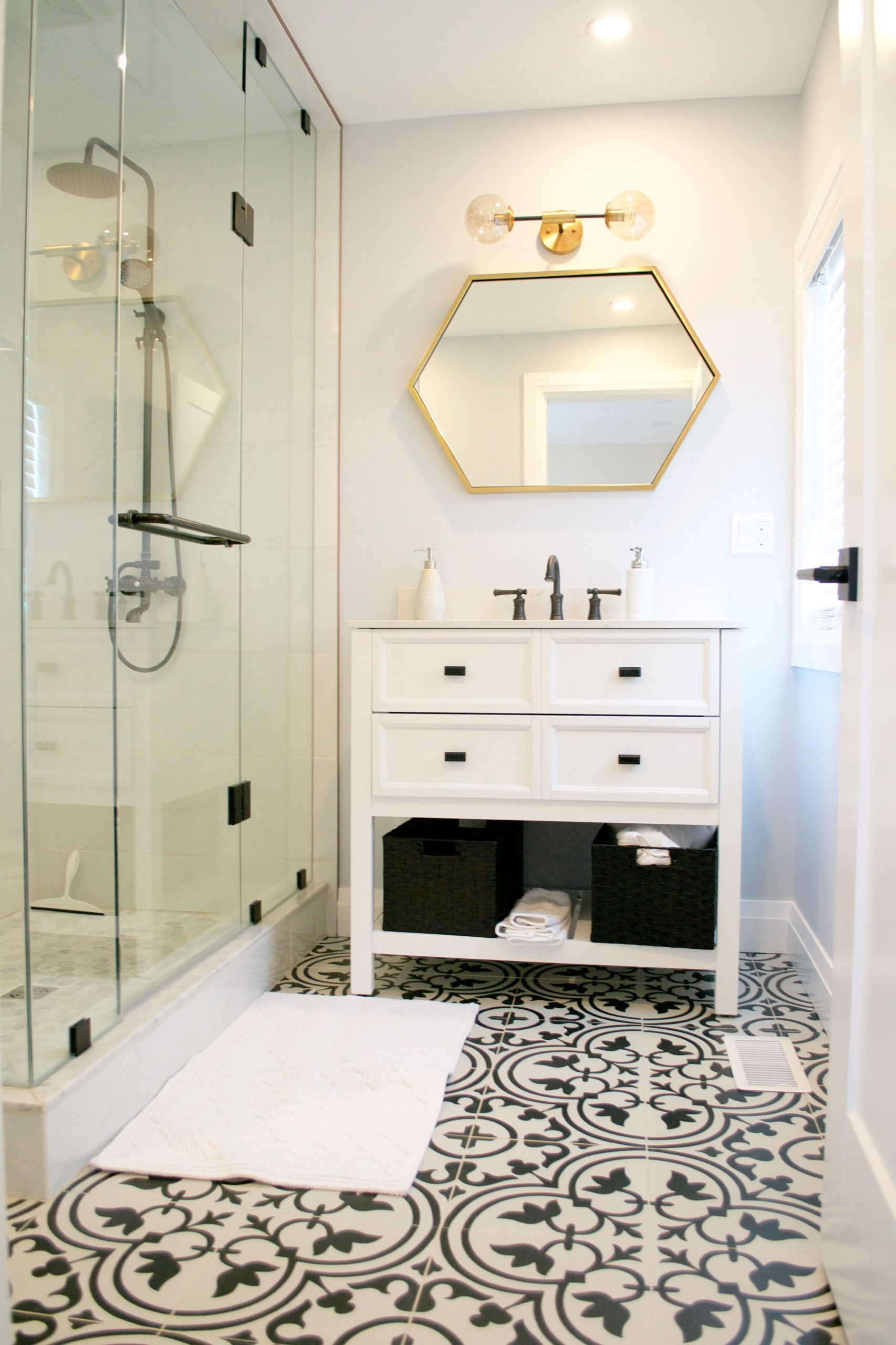 Bathroom with patterned floor, white vanity, glass shower, gold-framed mirror, and white walls.