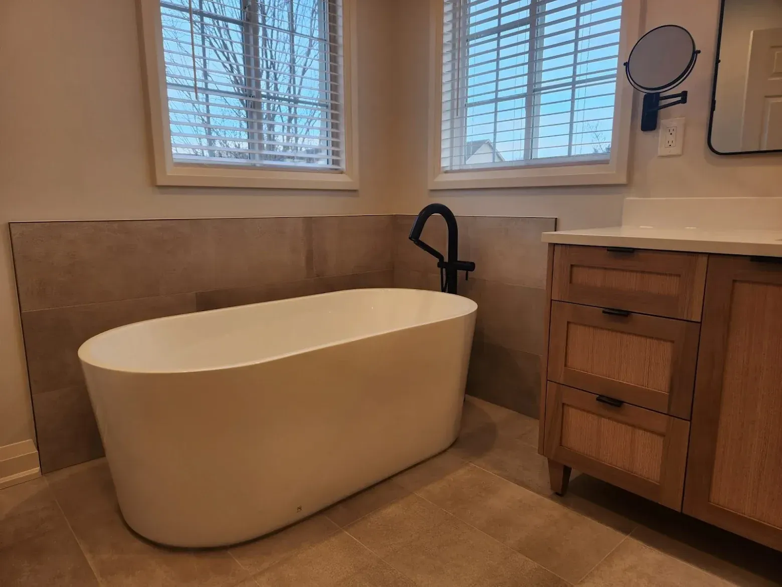 Freestanding white bathtub in a neutral-toned bathroom with two windows, and wood vanity.