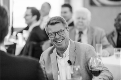 A man in a suit and glasses is sitting at a table with a glass of wine.