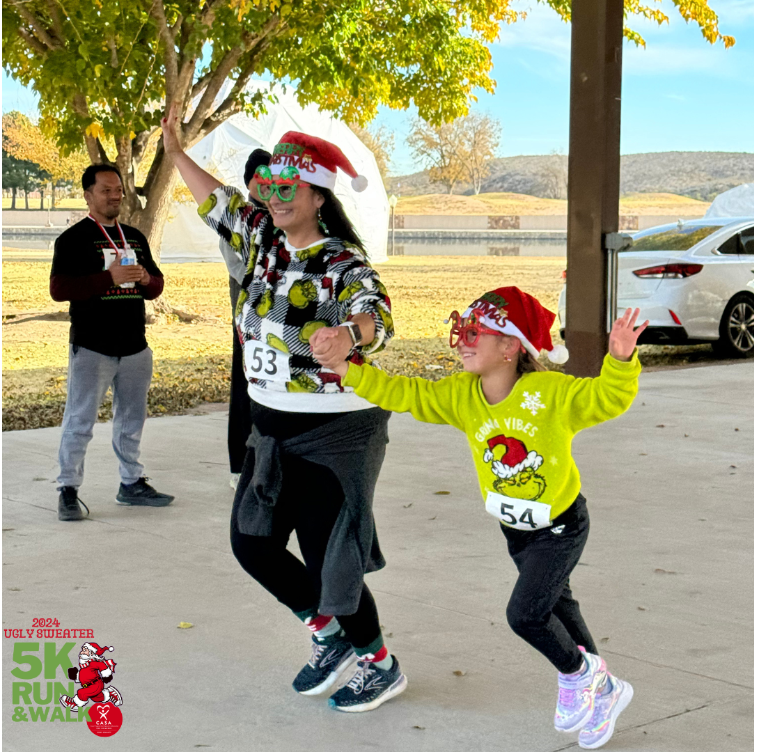 Two runners in festive hats race under trees while a man watches nearby at a community event