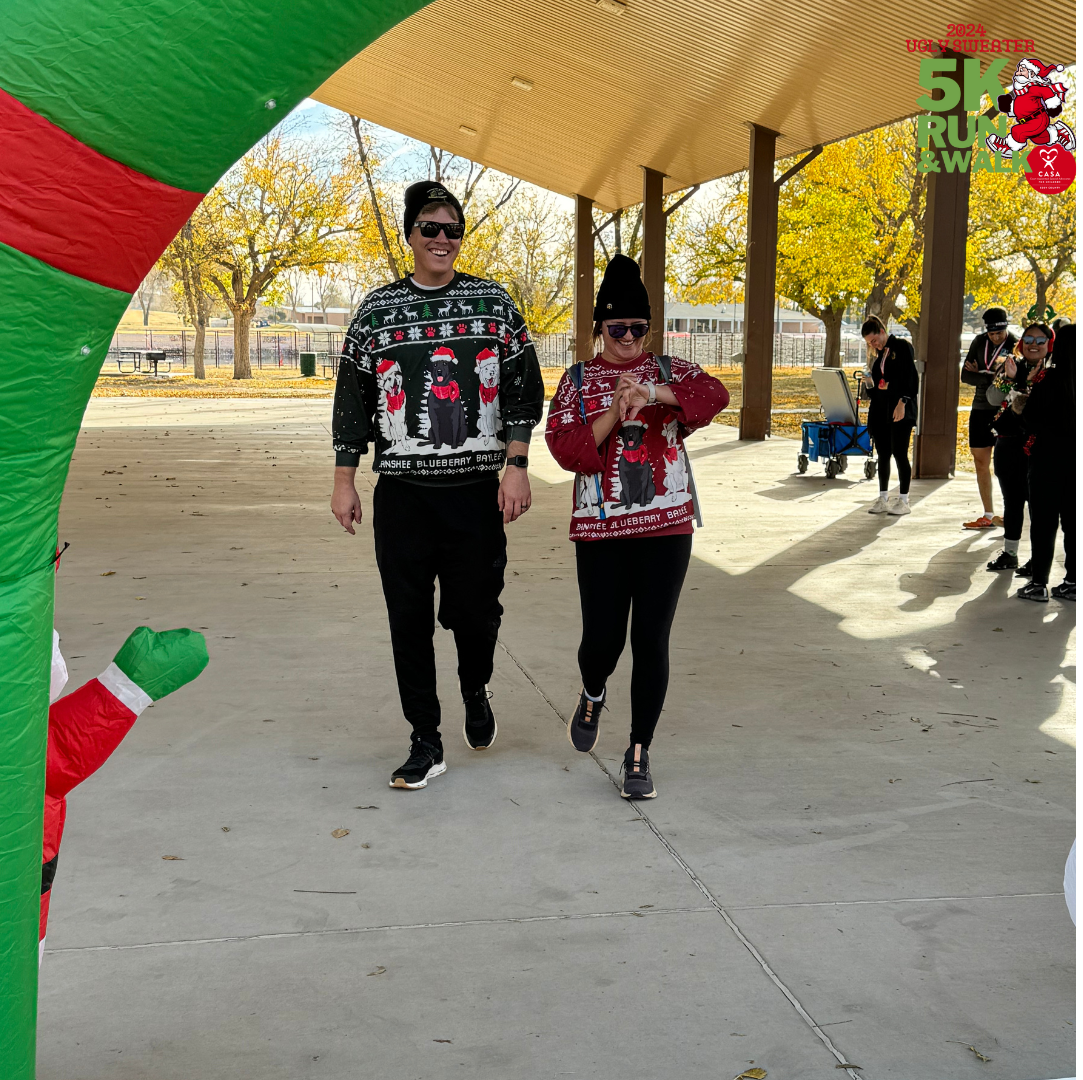 Two people in holiday sweaters walking under a decorated outdoor canopy in a park.