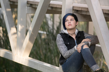 Person in a black vest sits on a wooden structure, resting one arm on a knee and looking upward.