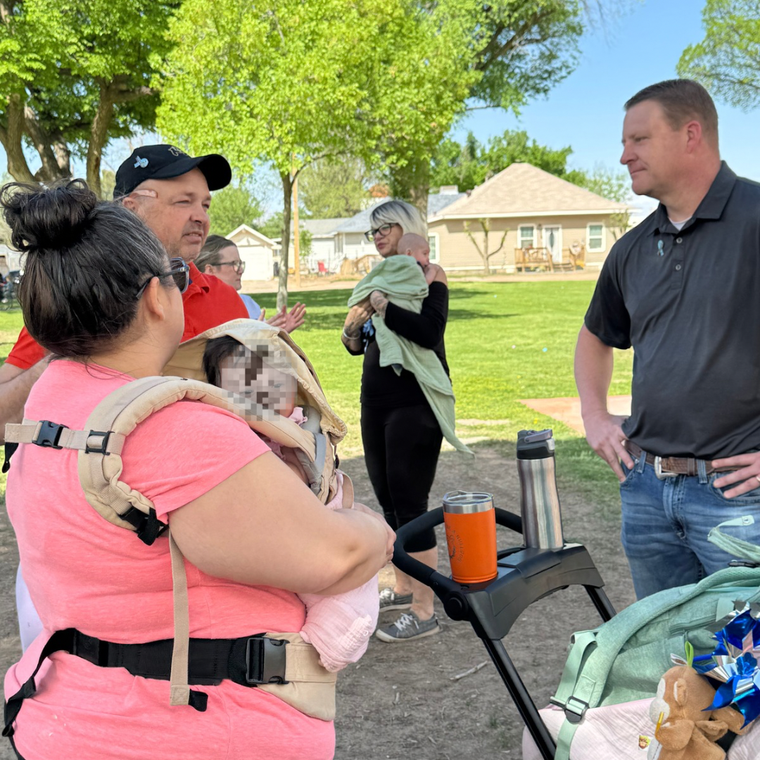 People chatting at a park picnic table, with a woman holding a small dog and a stroller nearby.