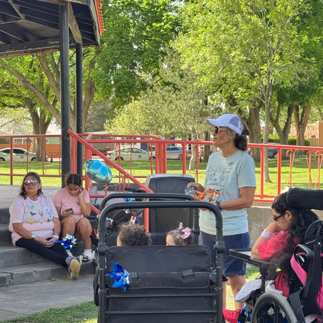 Woman speaking to seated group in a park, near a stroller under a shaded pavilion