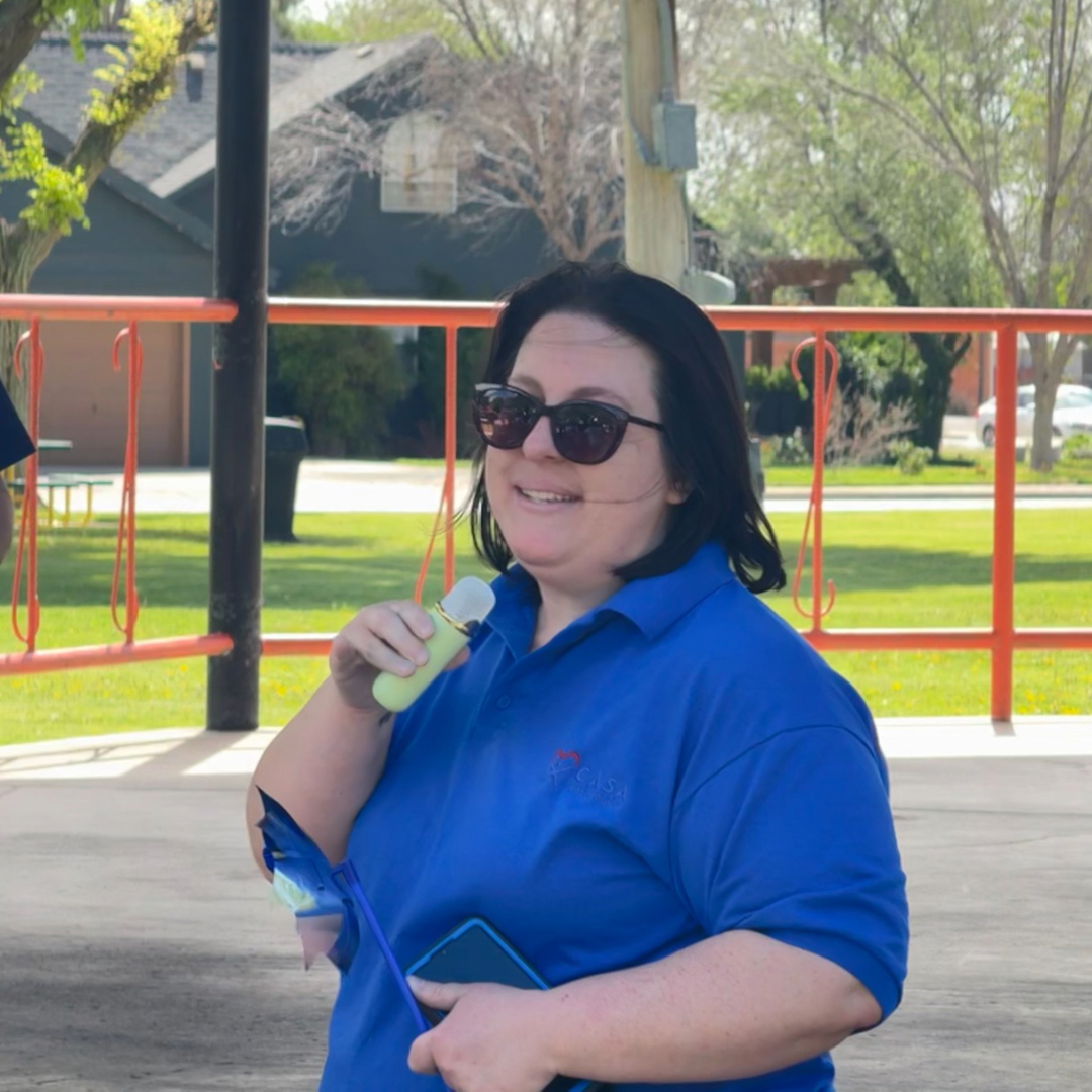 Woman in blue shirt and sunglasses holding a phone at a park pavilion with orange railings