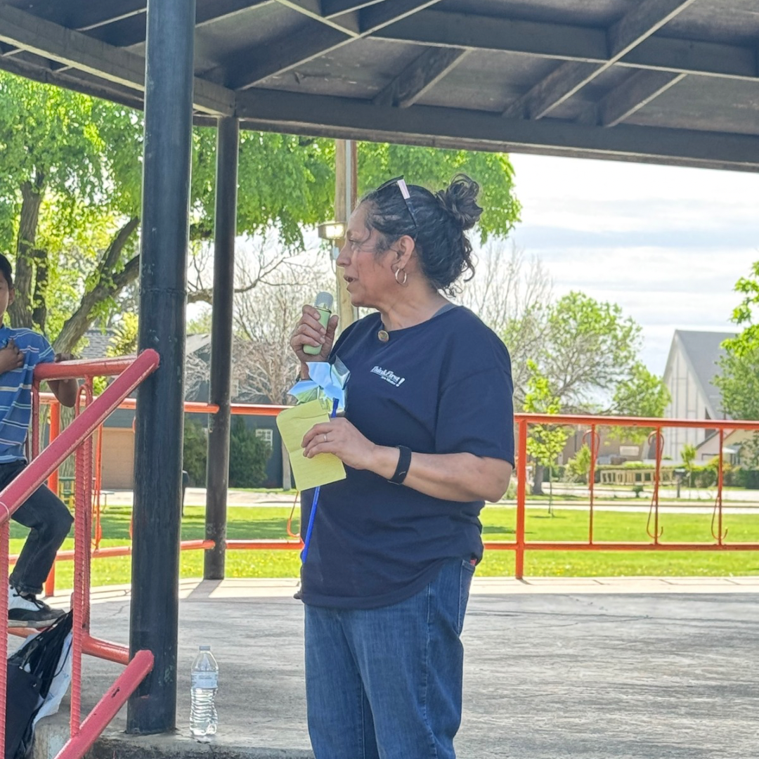 Woman in a navy shirt stands under a pavilion, holding a drink and looking left in a park.